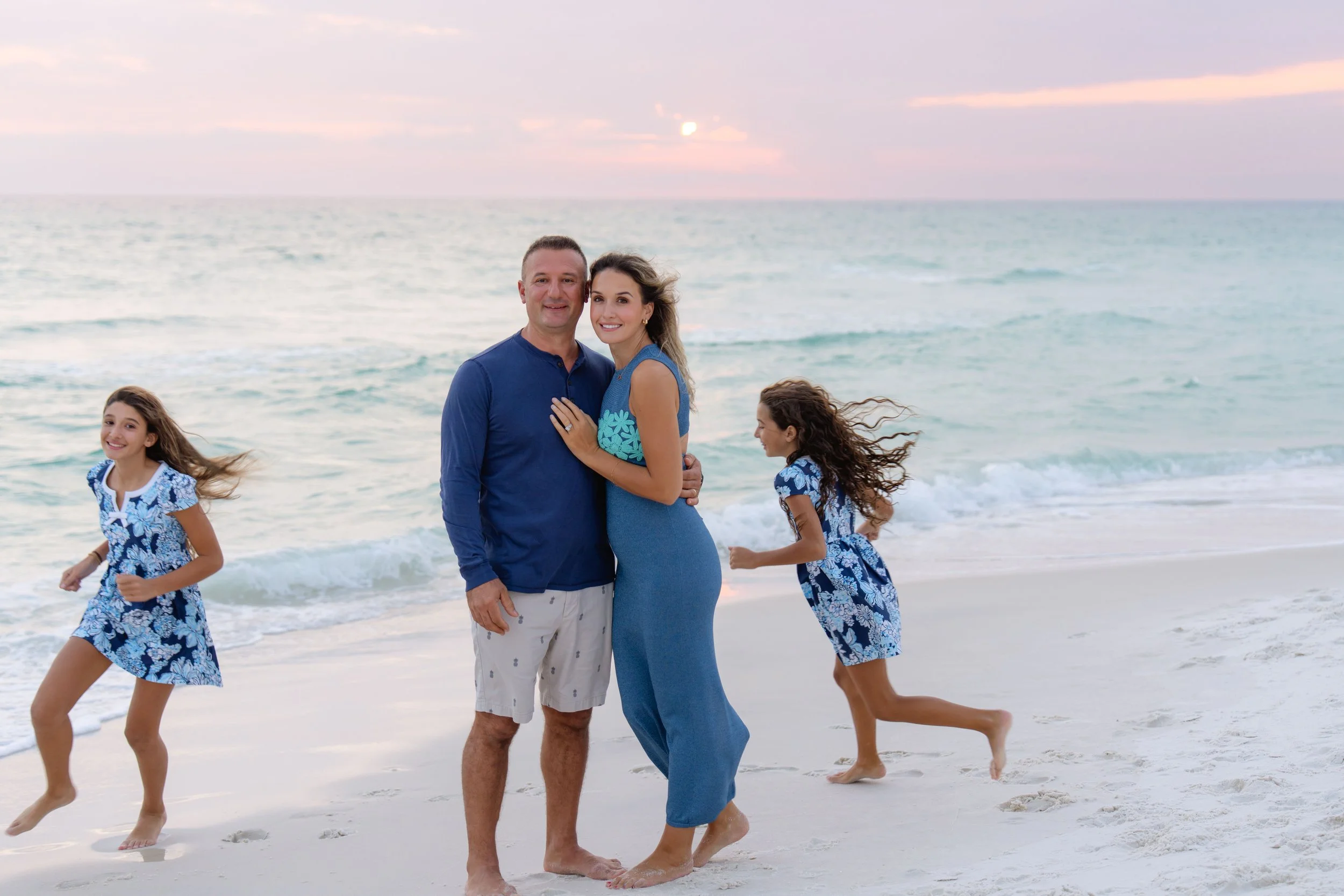 A family standing on a beach at sunset, with ocean waves in the background. Two adults, a man and a woman, are close together smiling at the camera. Two young girls in matching blue and white dresses are playing nearby, one running and the other standing.