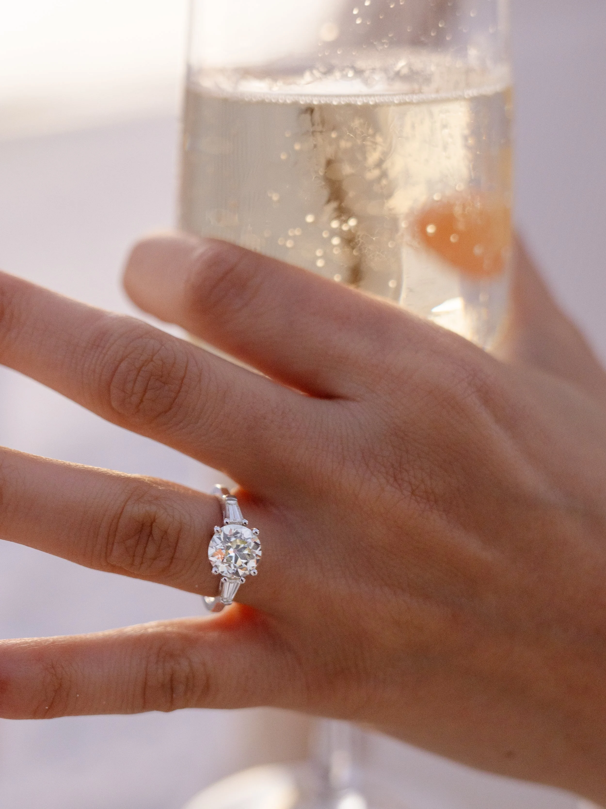 Woman holding glass of champagne showing engagement ring in the sunset.