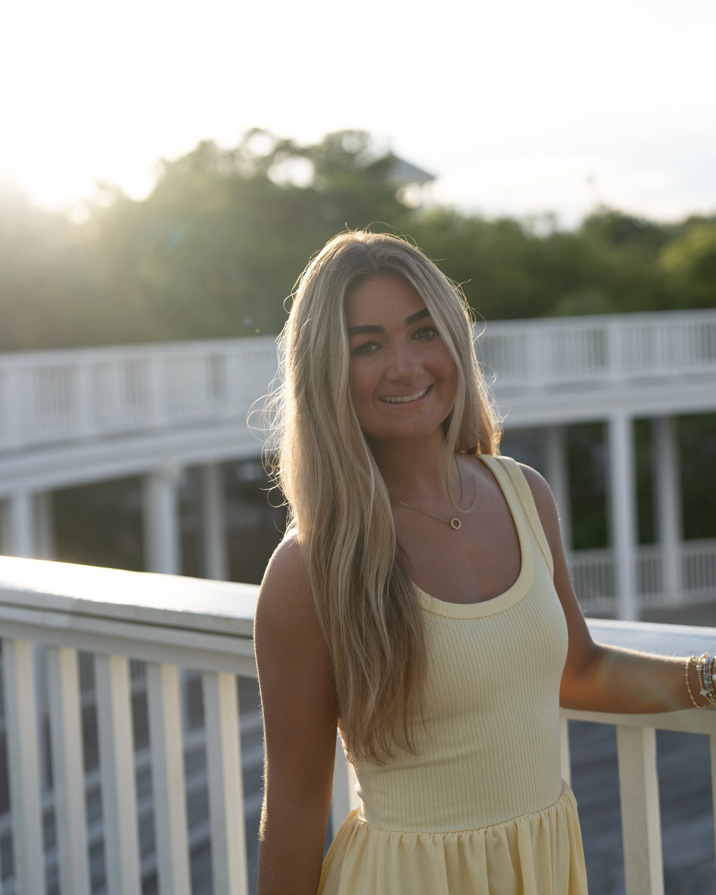 A young woman with long blonde hair smiling on a balcony during sunset, wearing a yellow sleeveless dress and jewelry.
