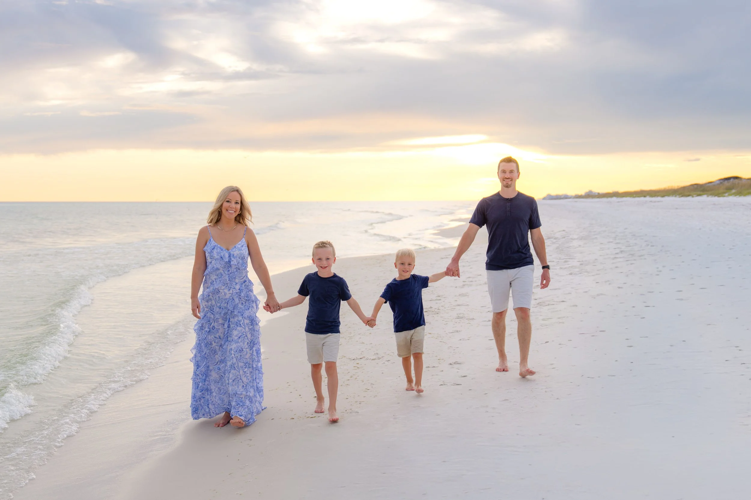 Family of four walking hand in hand on the beach at sunset, smiling, with ocean and cloudy sky in the background.