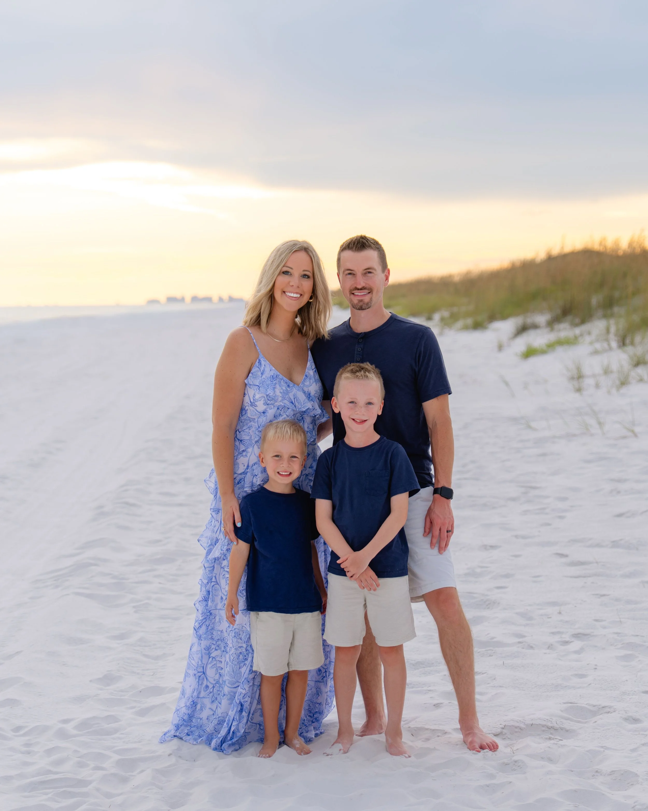 A family of four standing on a sandy beach at sunset, smiling at the camera. The mother and father are standing behind two young boys, all dressed in casual summer clothes.