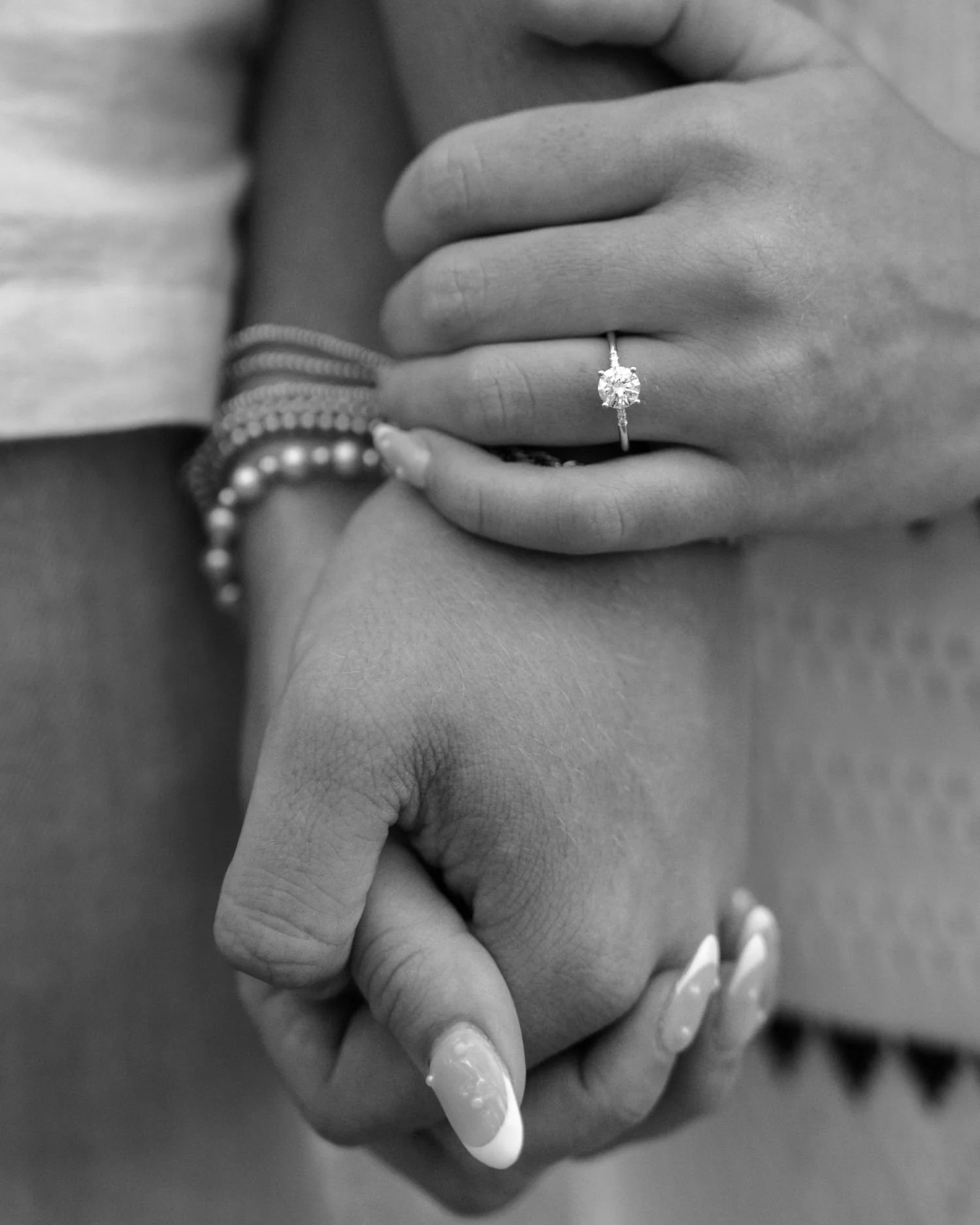 Close-up of two hands clasped together, one hand with an engagement ring featuring a large gemstone, both hands adorned with additional jewelry, in black and white.