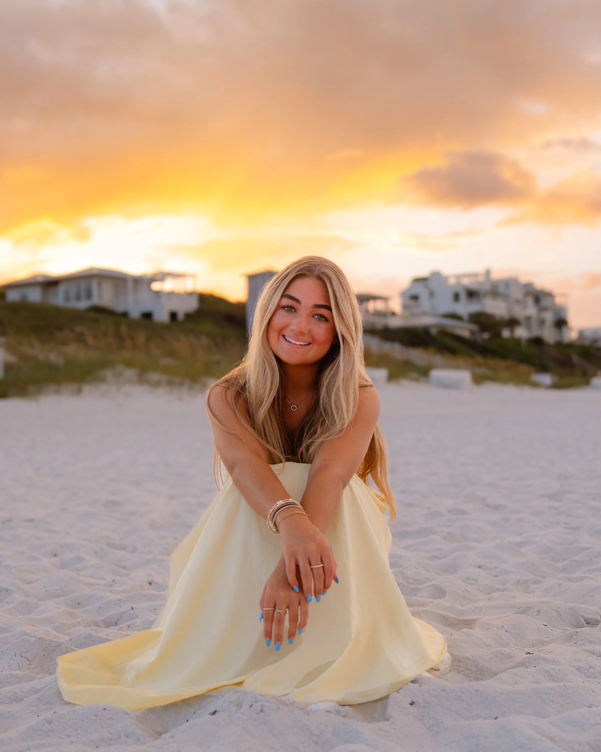 A young woman with long blonde hair sitting on the sand at the beach during sunset, smiling at the camera, wearing a yellow dress and jewelry with houses in the background.