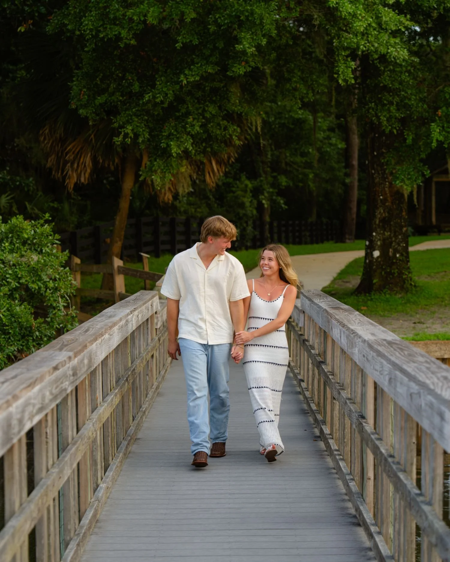 A young couple walking hand in hand on a wooden bridge, smiling at each other, surrounded by green trees in a park.
