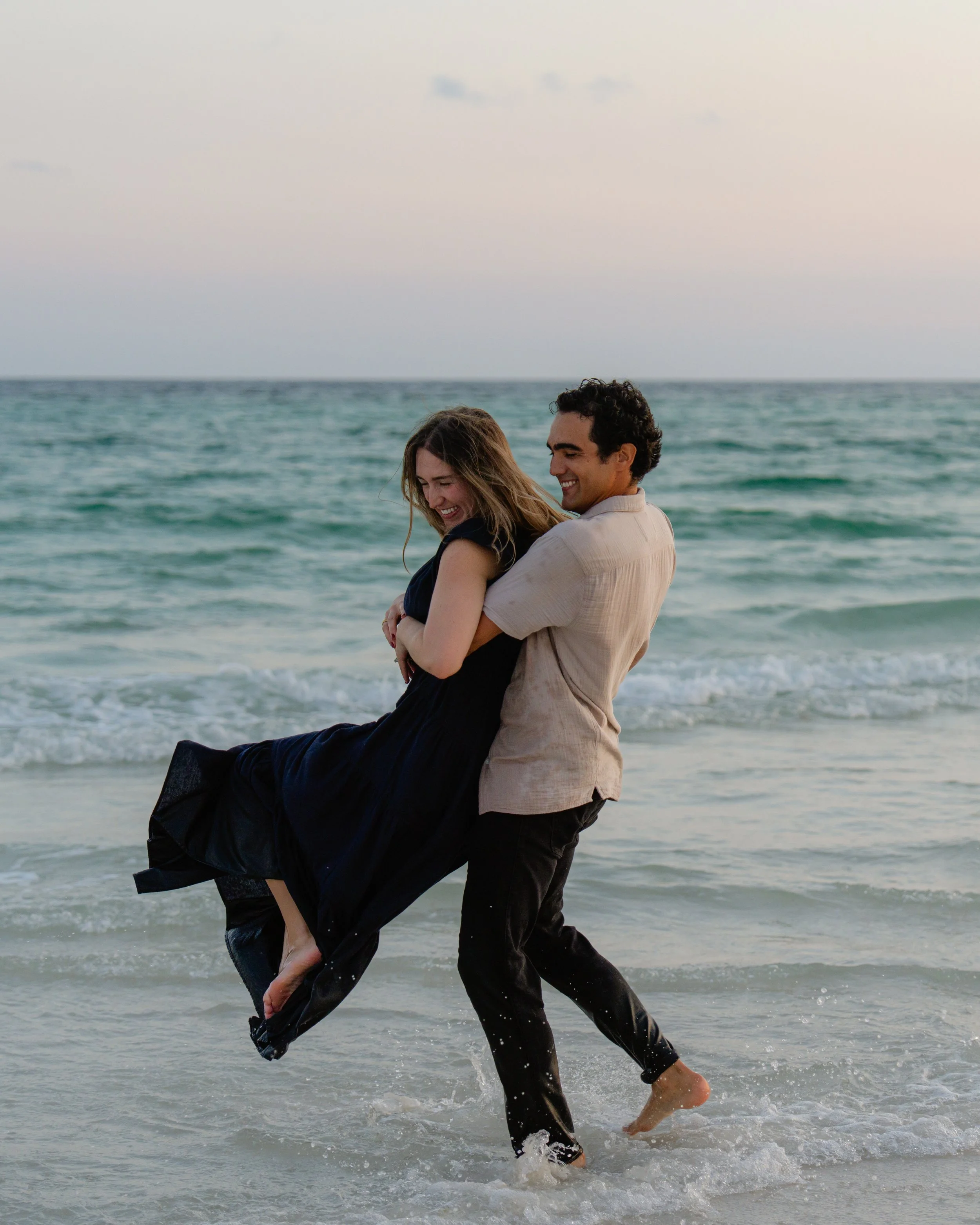 A man lifting a woman in the water at the beach during sunset, both smiling