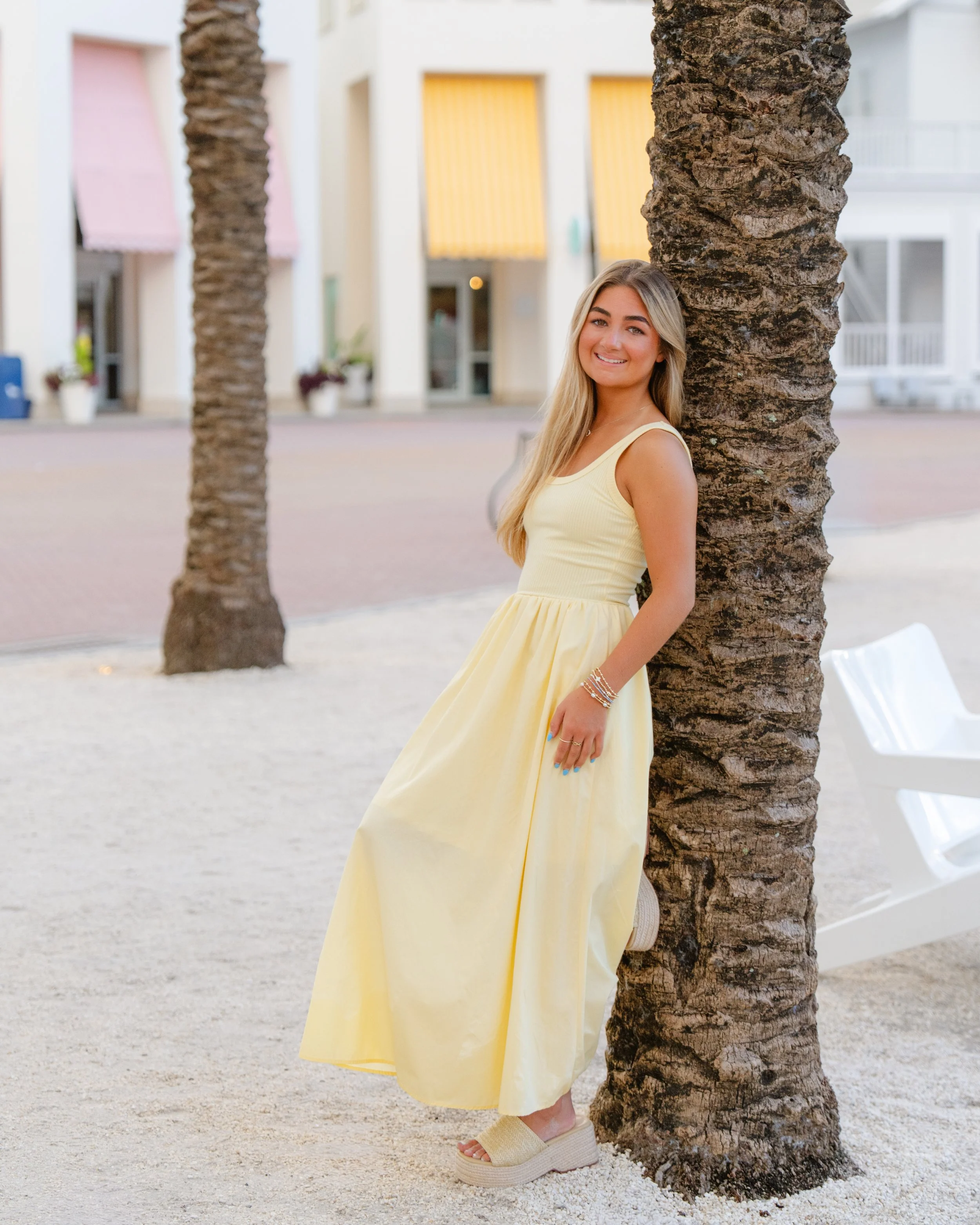 A young woman in a yellow dress leaning against a palm tree on a sandy area with buildings in the background.