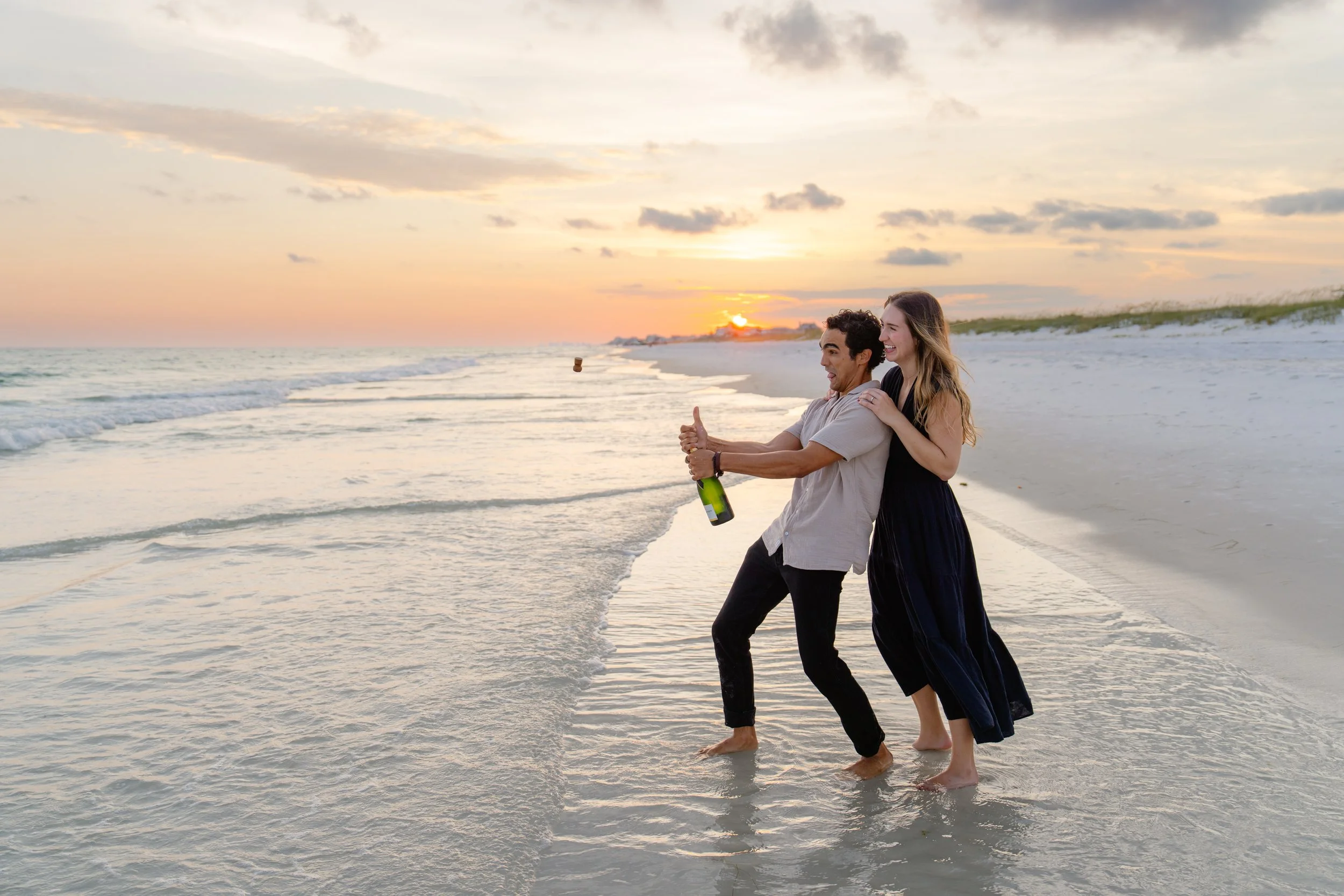 A happy couple on a beach at sunset, with the man opening a bottle of champagne while the woman embraces him from behind.