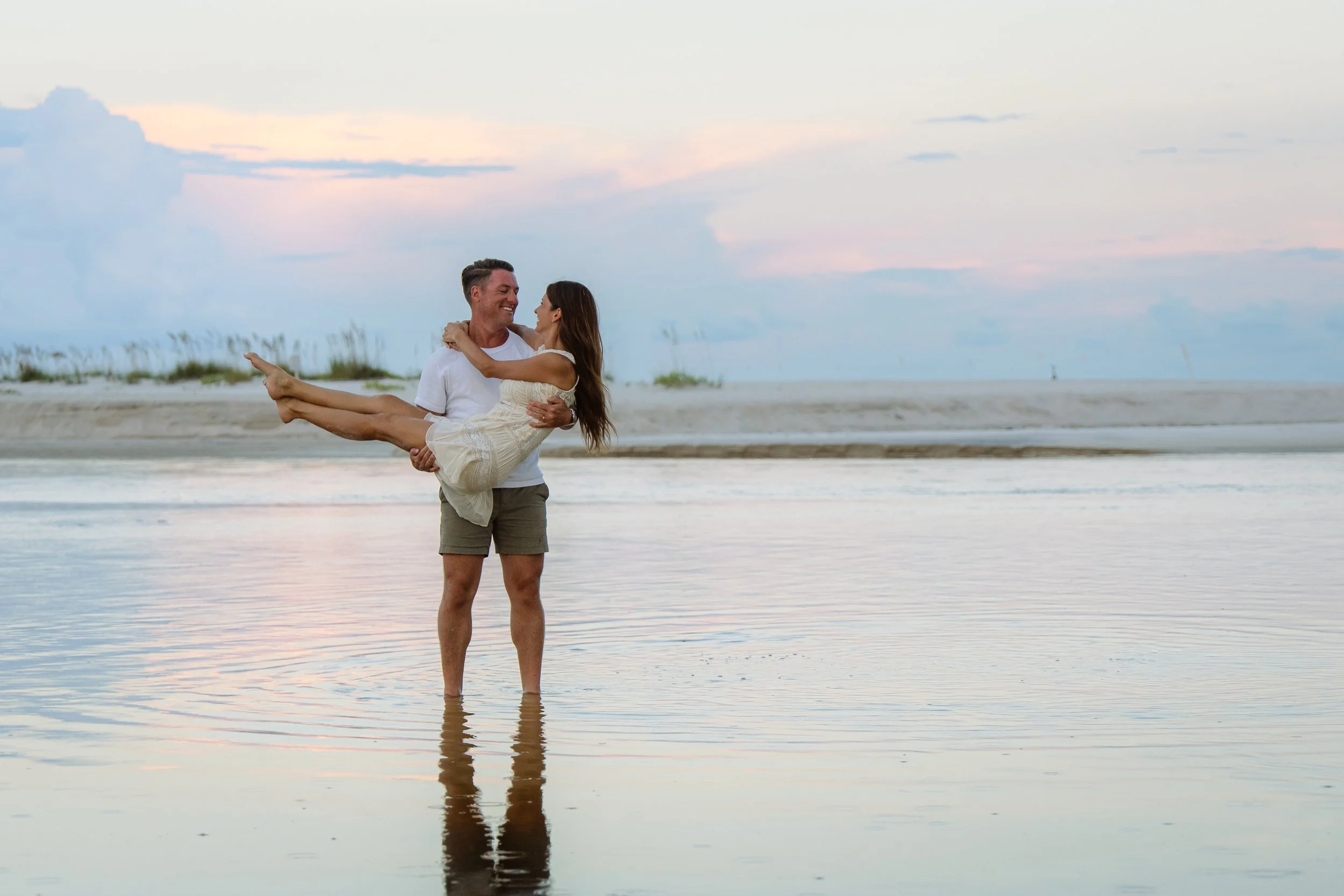 A man holding a woman in his arms at the beach during sunset.