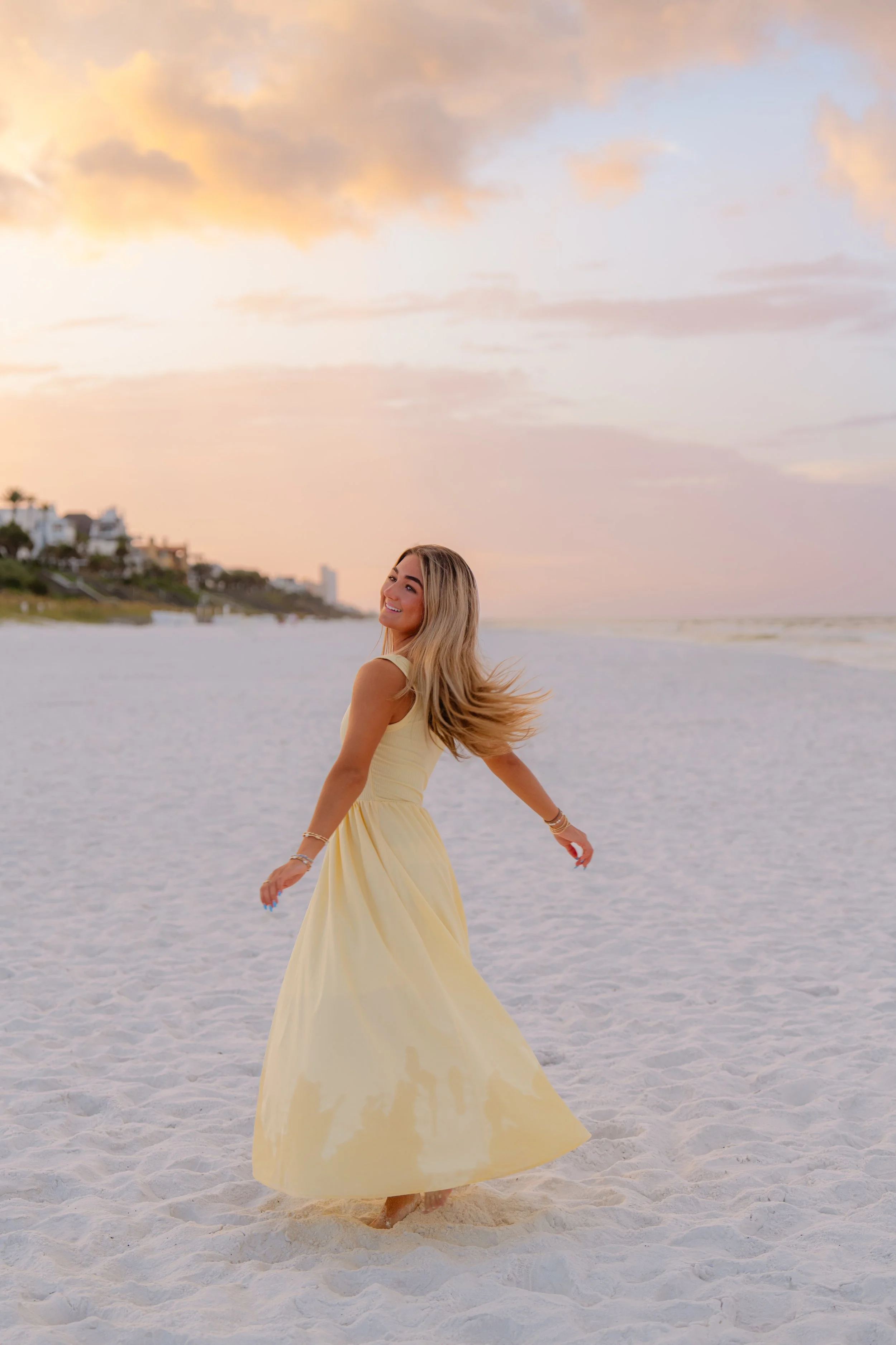 A woman in a yellow dress spinning on a sandy beach at sunset, with pastel-colored sky and clouds, and houses visible in the background.