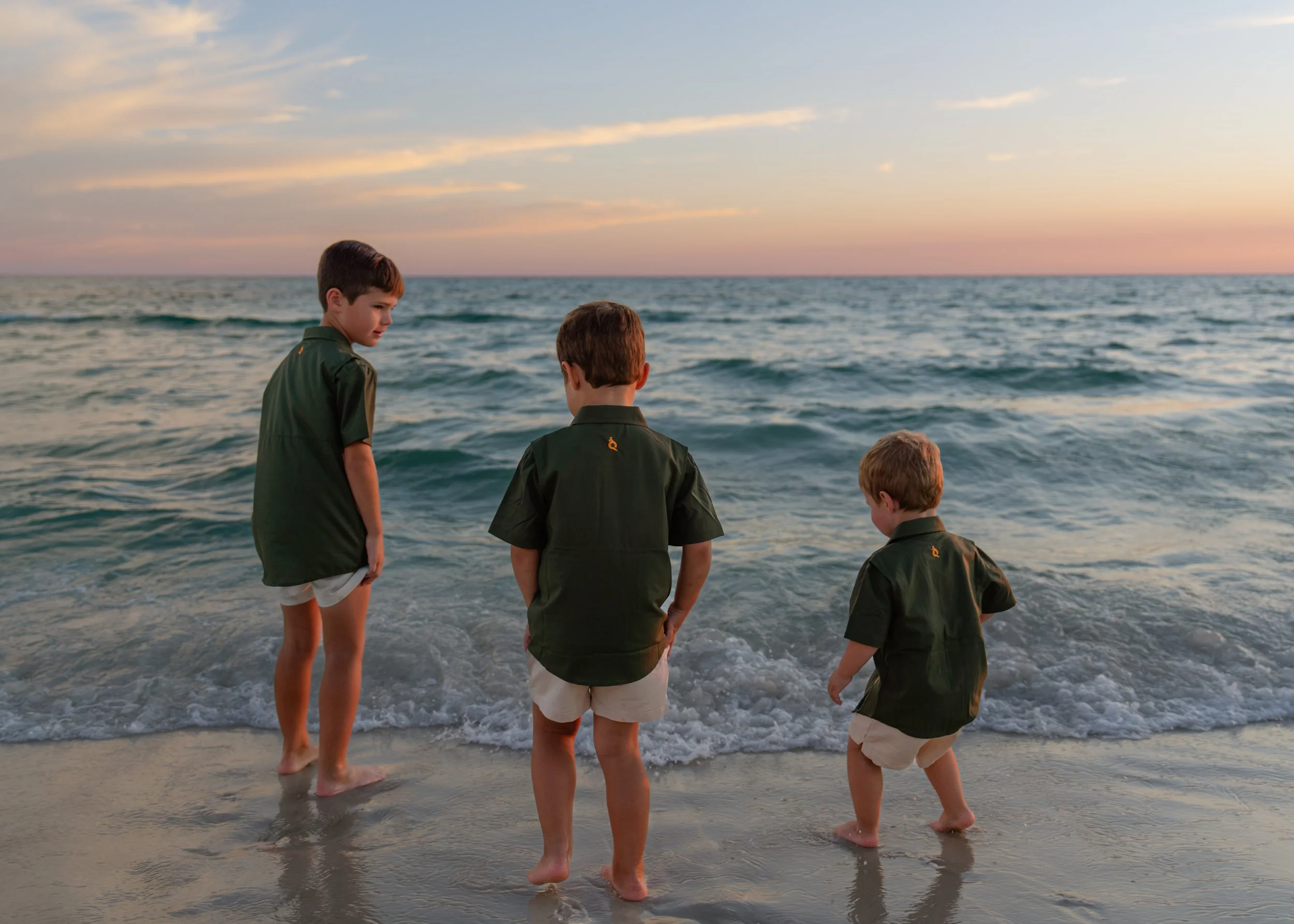 Four boys in green shirts and white shorts stand on a sandy beach at sunset, looking at the ocean waves.