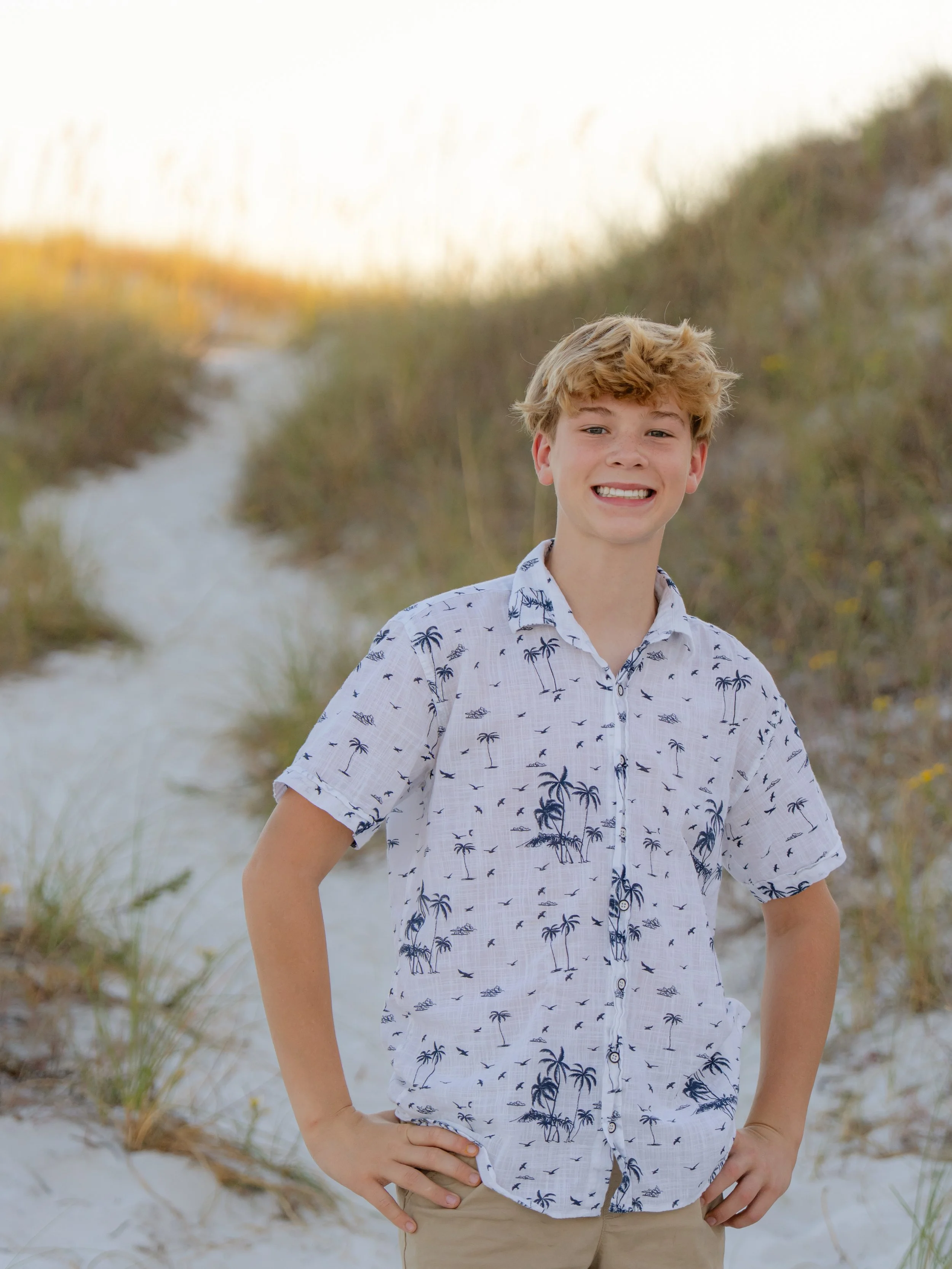 Young man with blonde hair in a white shirt standing with his hands on his hips with the dunes behind him.