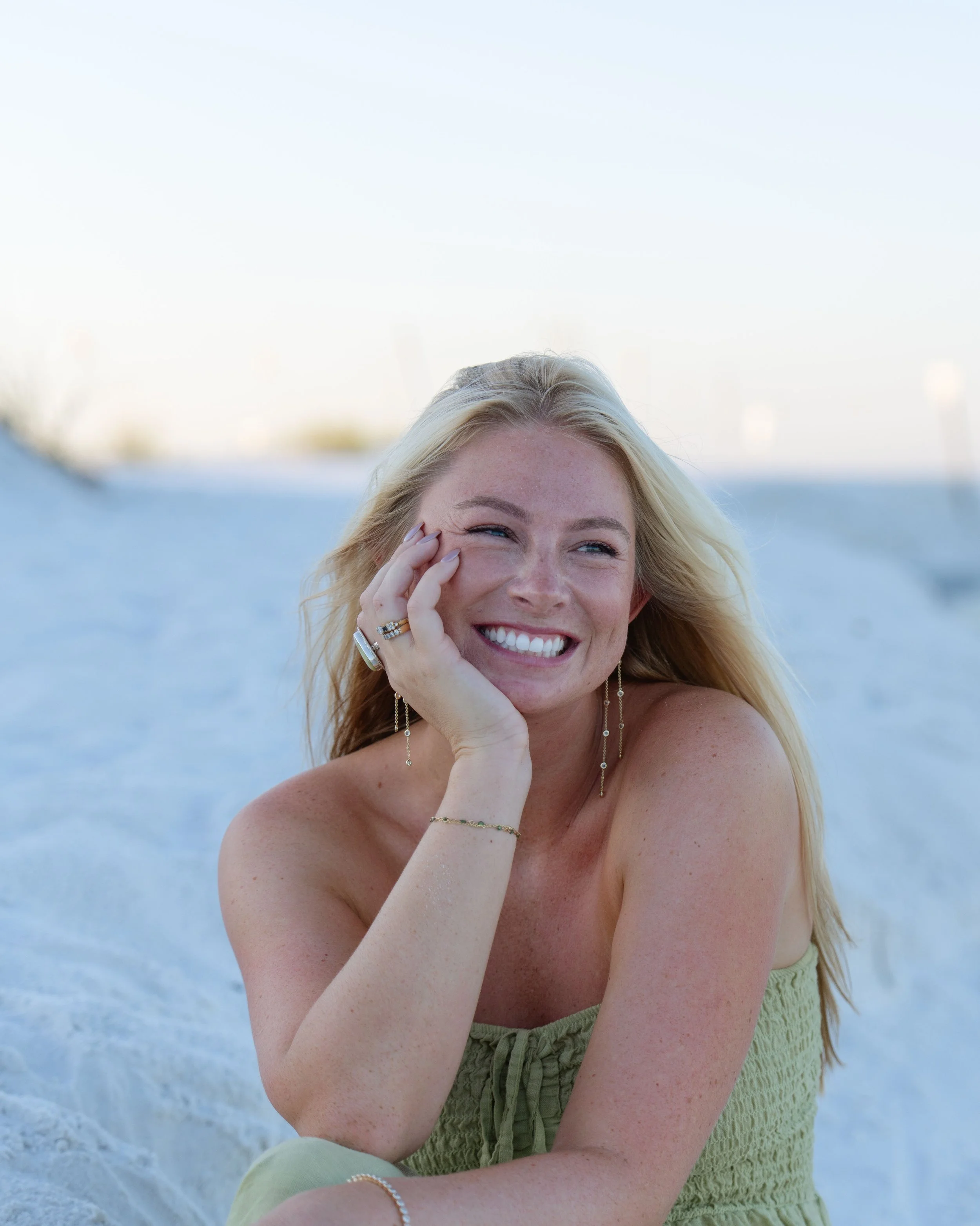 A smiling woman with blonde hair sitting on a sandy beach, wearing jewelry, in daylight.
