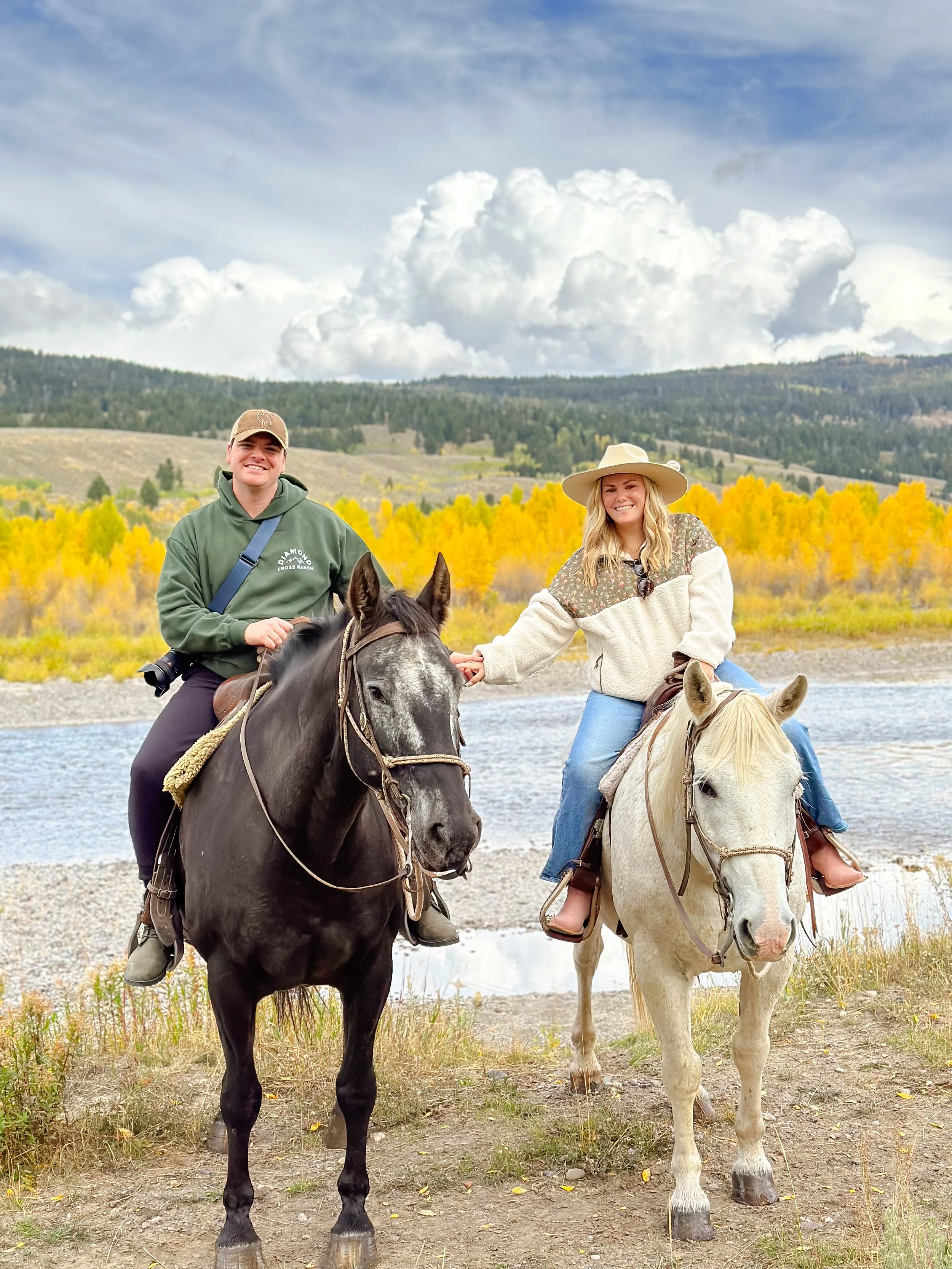 Two women riding horses by a river, with yellow trees and hills in the background under a cloudy sky.