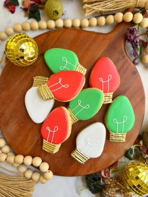 Decorative holiday cookies shaped like Christmas lights in red, green, and white colors with gold and white icing details, placed on a wooden serving board.