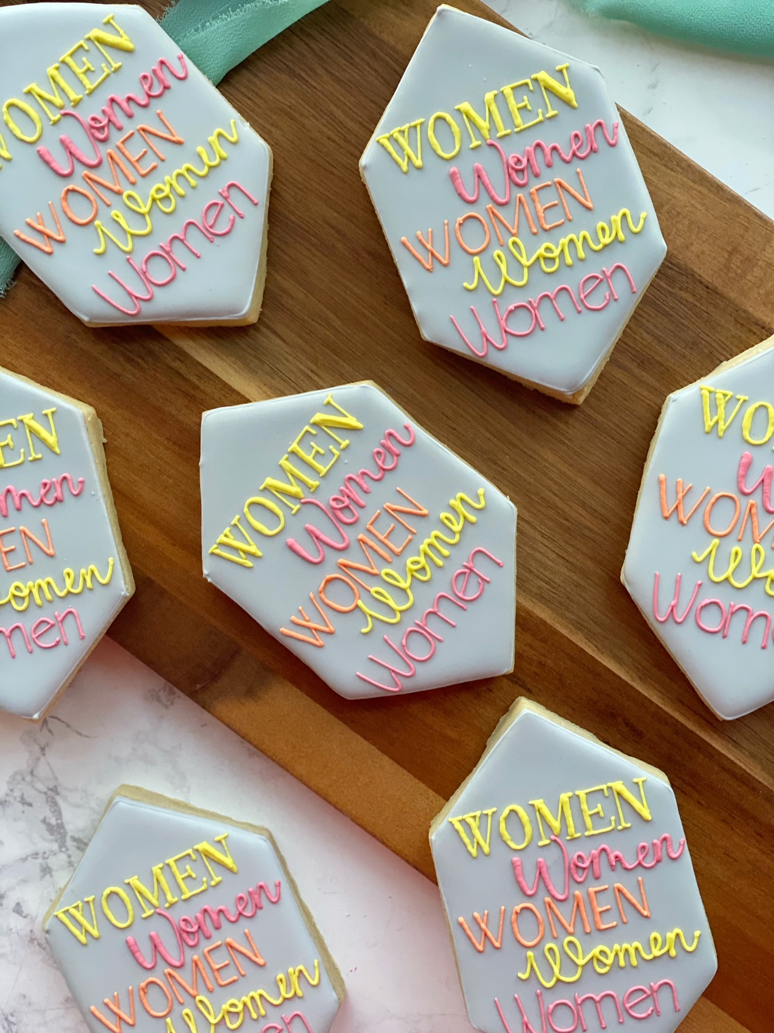 Decorative sugar cookies with white icing and colorful piped words 'WOMEN' in yellow, pink, orange, and yellow icing. The cookies are on a wooden surface and there is a green napkin in the background.