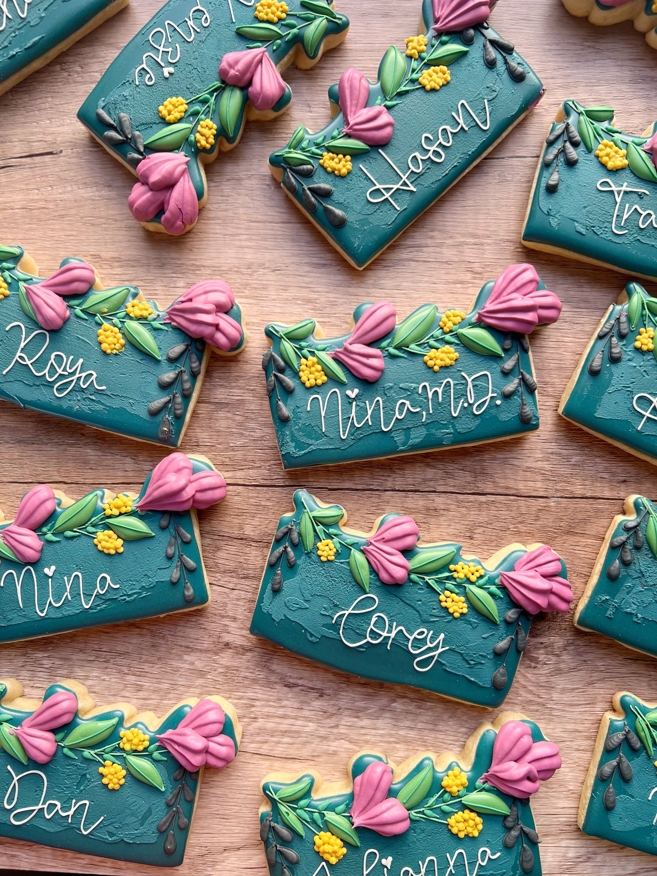 Decorated cookies with blue icing, pink flowers, green leaves, and yellow accents, personalized with names like Nina, Tory, and Lasaw.