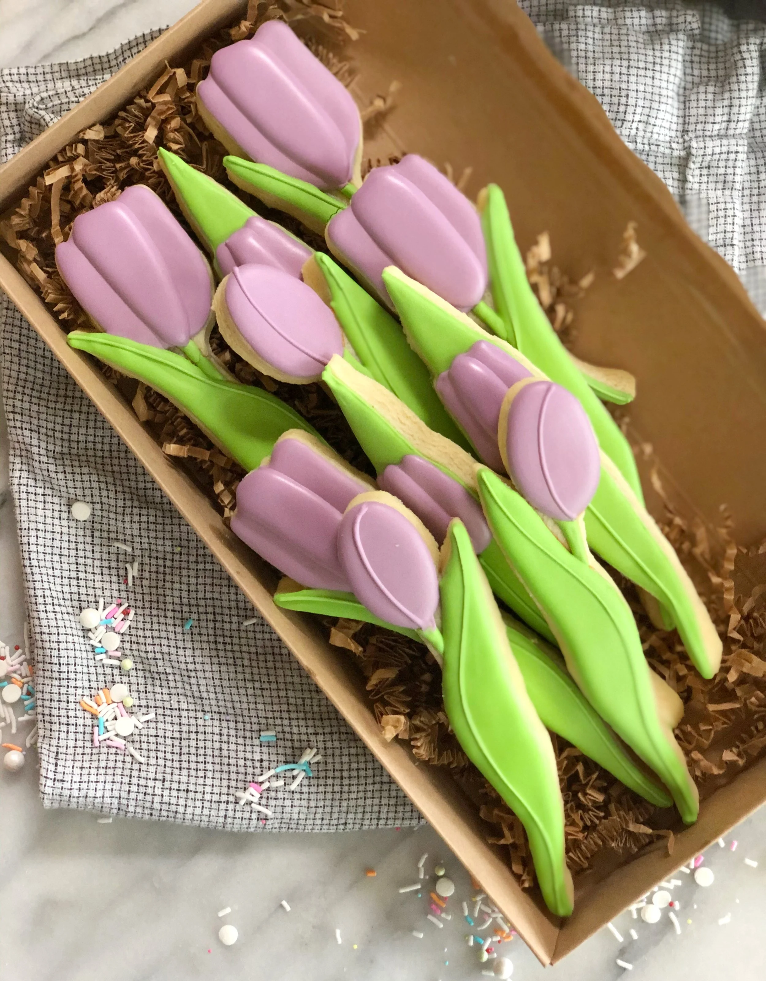 A box of flower-shaped sugar cookies with pink icing and green stems, surrounded by rainbow-colored sprinkles on a light surface with a checked cloth.