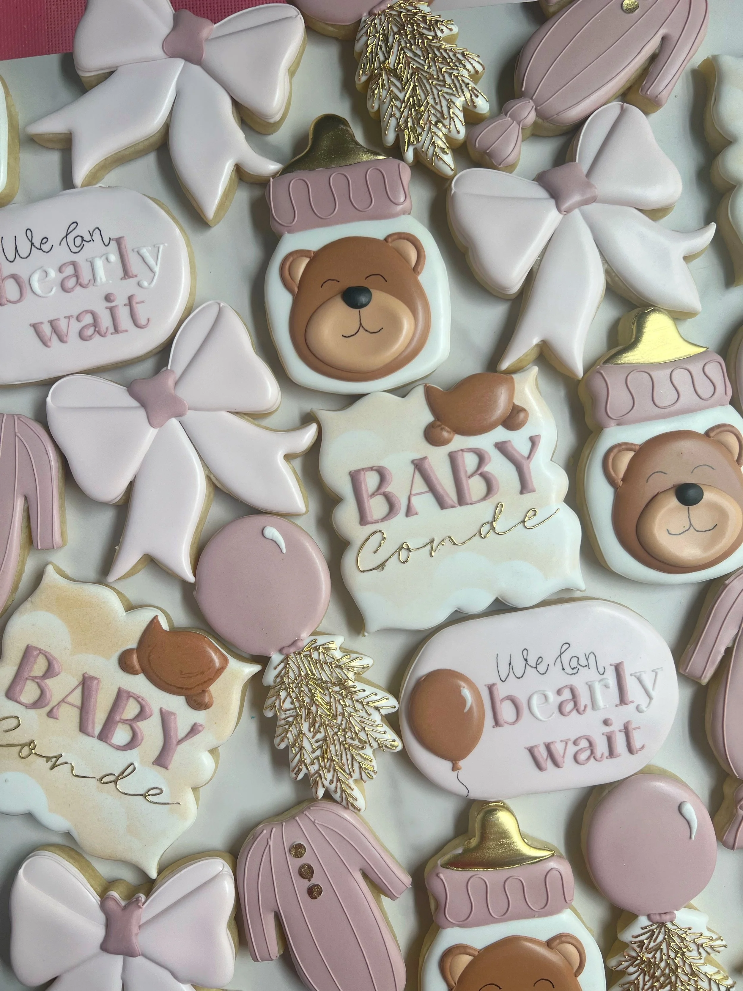 A collection of decorated cookies with baby-themed designs, including teddy bears, baby bottles, bows, and leaves, in pastel pink, white, and gold colors.
