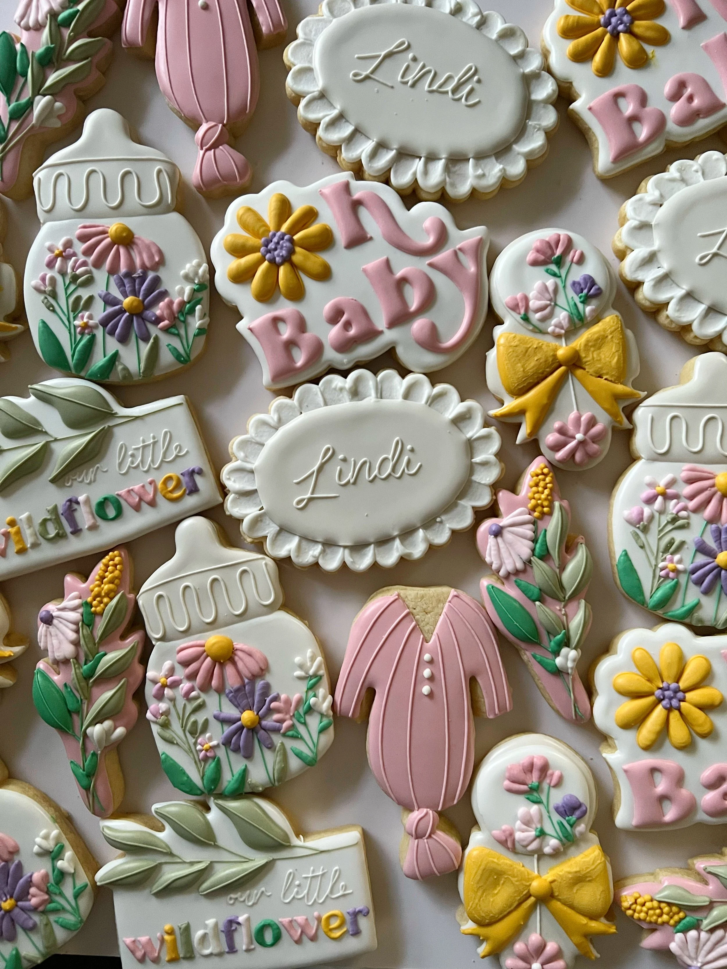 Decorated cookies with floral and baby shower themes, featuring names like 'Lindi' and 'Babi,' with pastel pink, yellow, purple, and white icing.