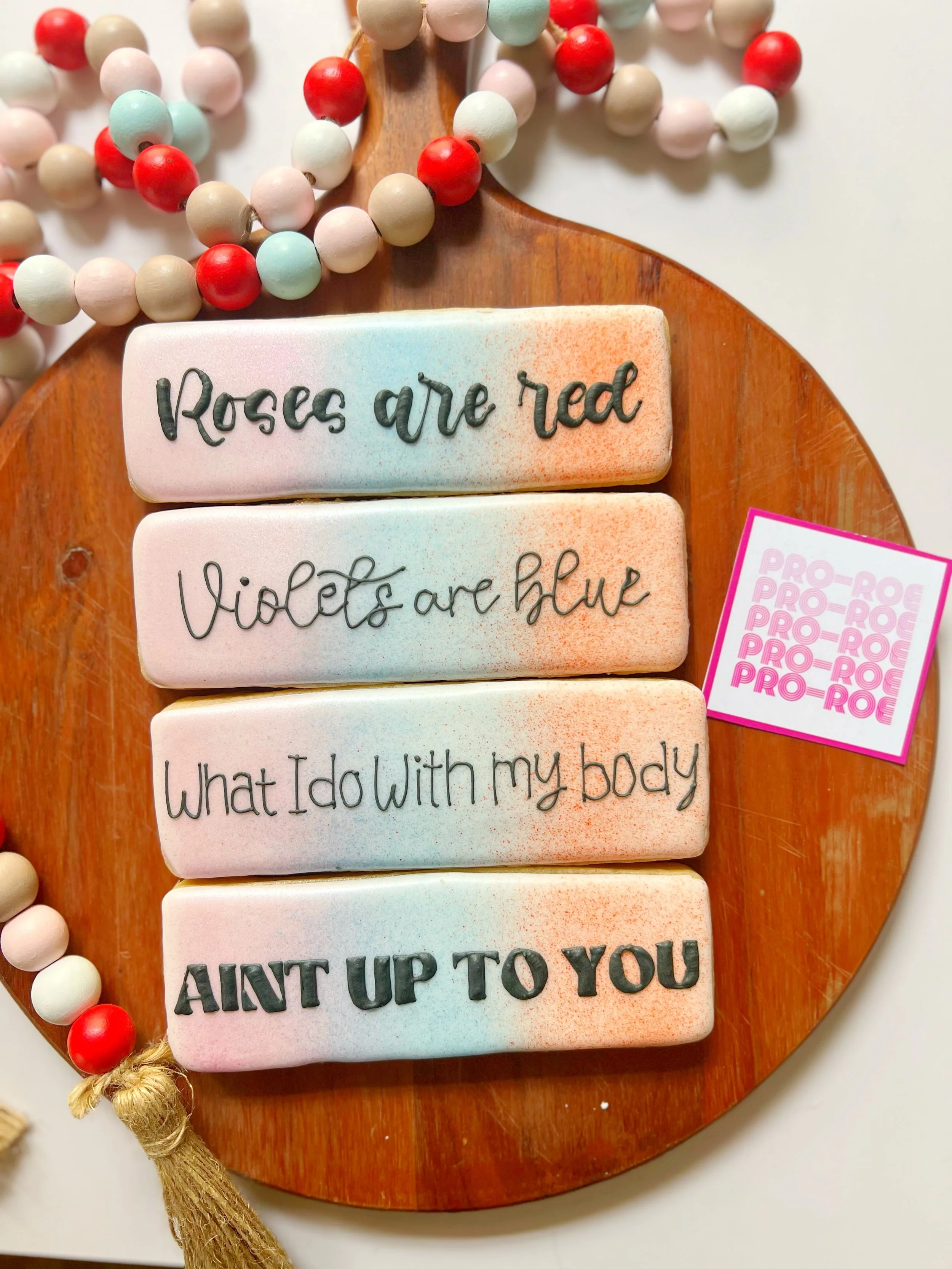 Colorful clay tiles with handwritten phrases about roses, violets, and personal sentiments, arranged on a wooden tray with a beaded necklace and a pink sticker nearby.