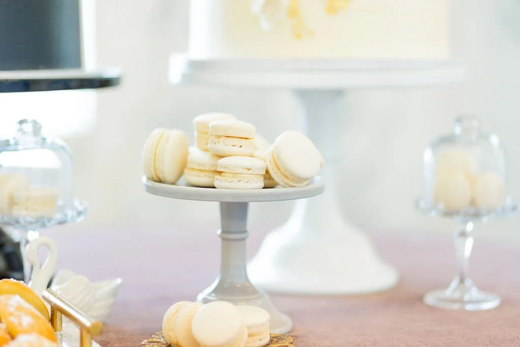 White macarons arranged on a small white cake stand with a blurred dessert display in the background.