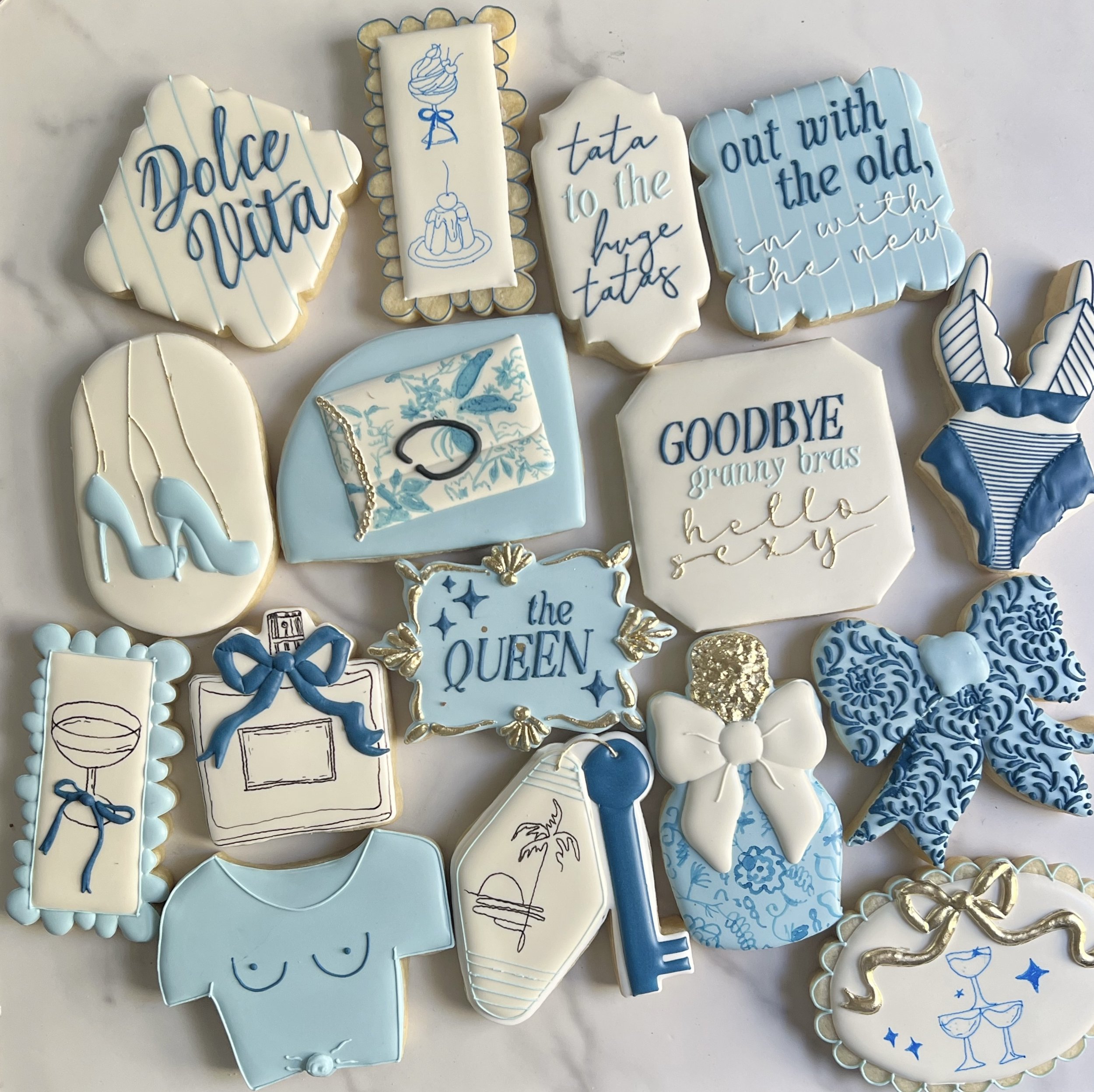 Collection of decorated cookies with various blue and white designs and messages, including cookies with phrases like "Dolce vita," "tata," "out with the old in with the new," and others featuring illustrations of desserts, drinks, and celebratory themes.