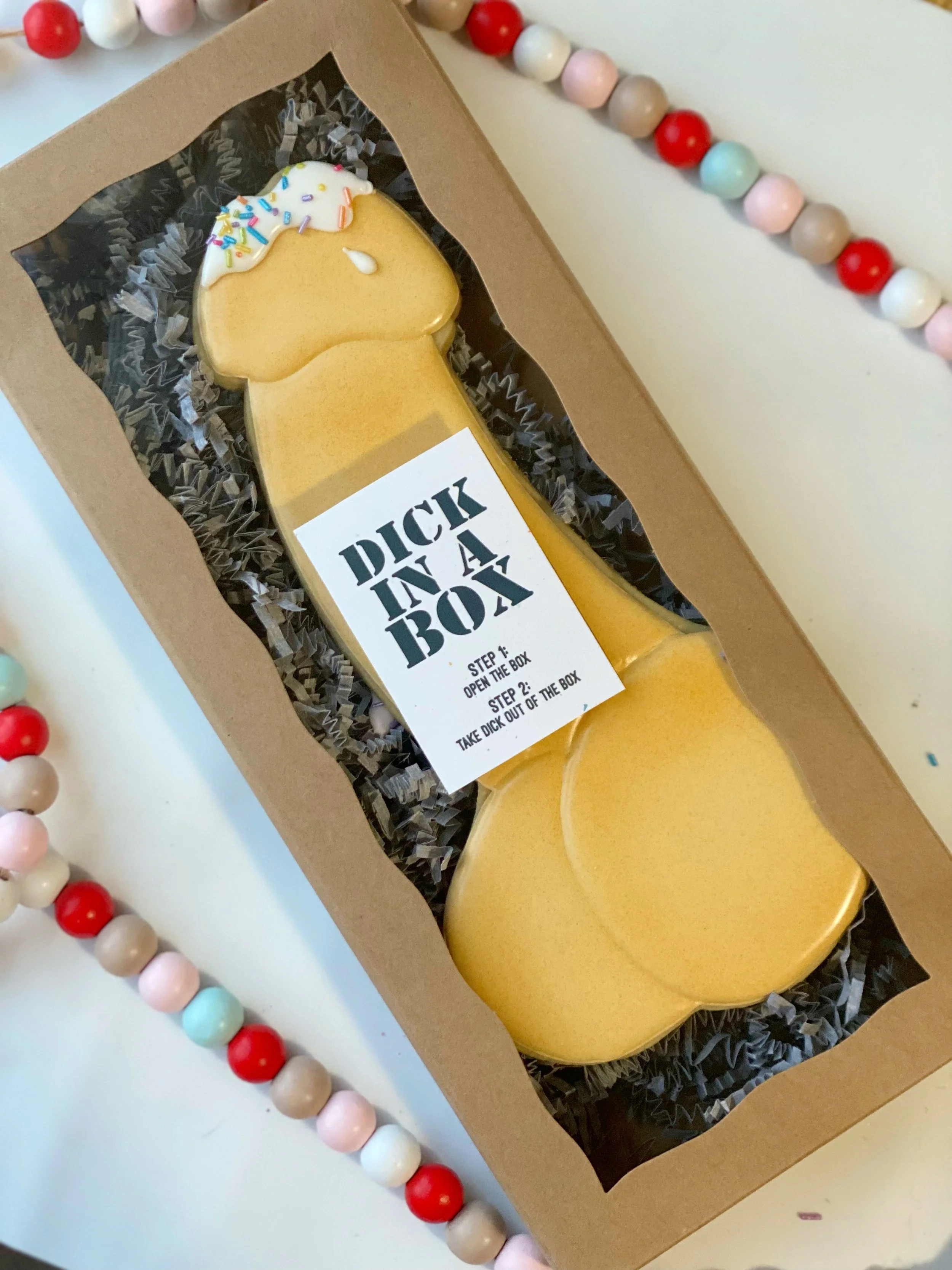 A box of yellow, ice cream-shaped cookies with white and colorful frosting and sprinkles, nestled in black shredded paper, with a label that reads 'DICK IN A BOX' and instructions to open the box and take out the cookie, surrounded by a bead garland with red, white, pink, turquoise, beige, and gray beads.
