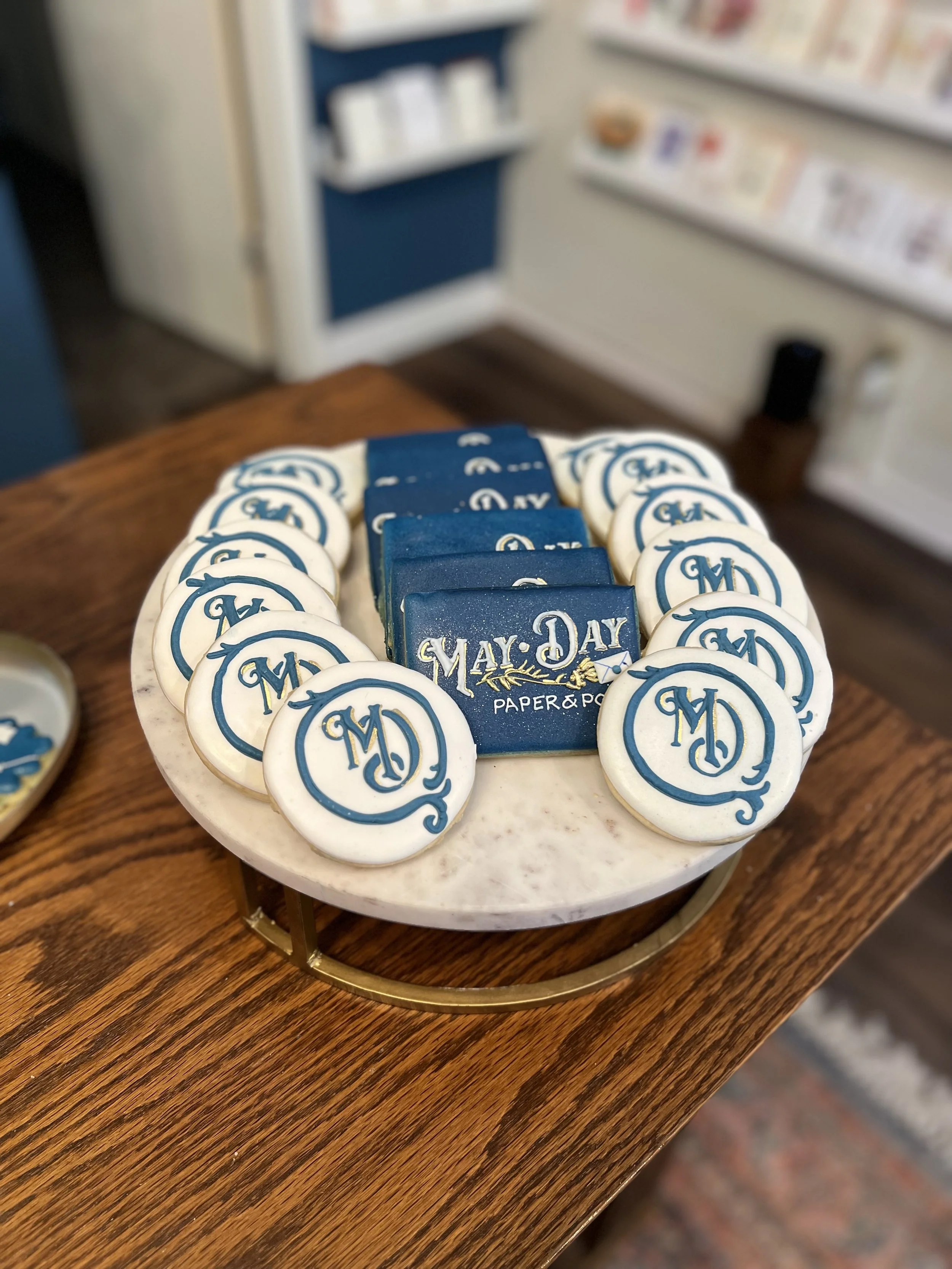 A round platter of decorated cookies with May Day theme, featuring the words "May Day" and blue monogrammed designs, on a wooden table with a blurred background of interior decor.