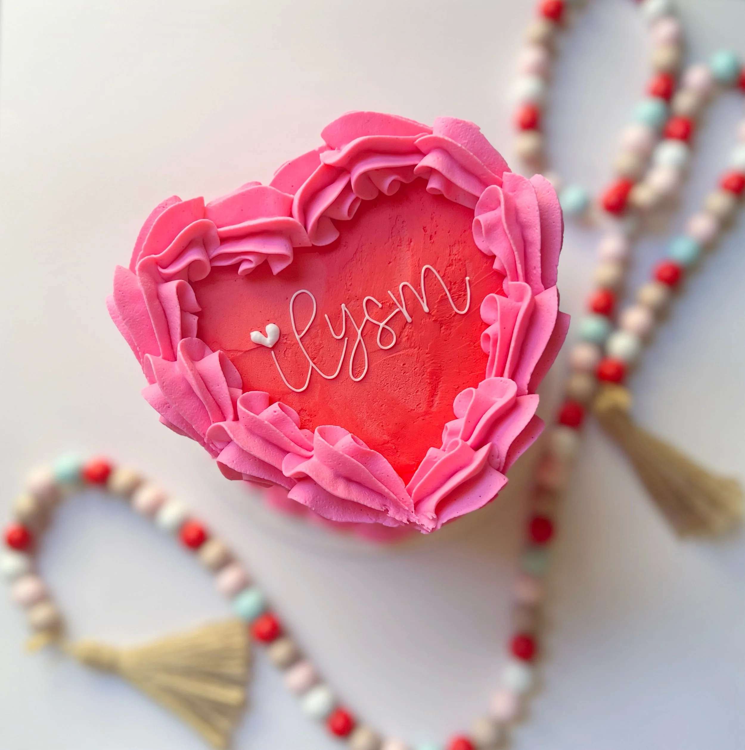 Close-up of a pink heart-shaped cookie decorated with pink frosting, with the word "love" written in white icing, and blurred multicolored bead necklaces in the background.