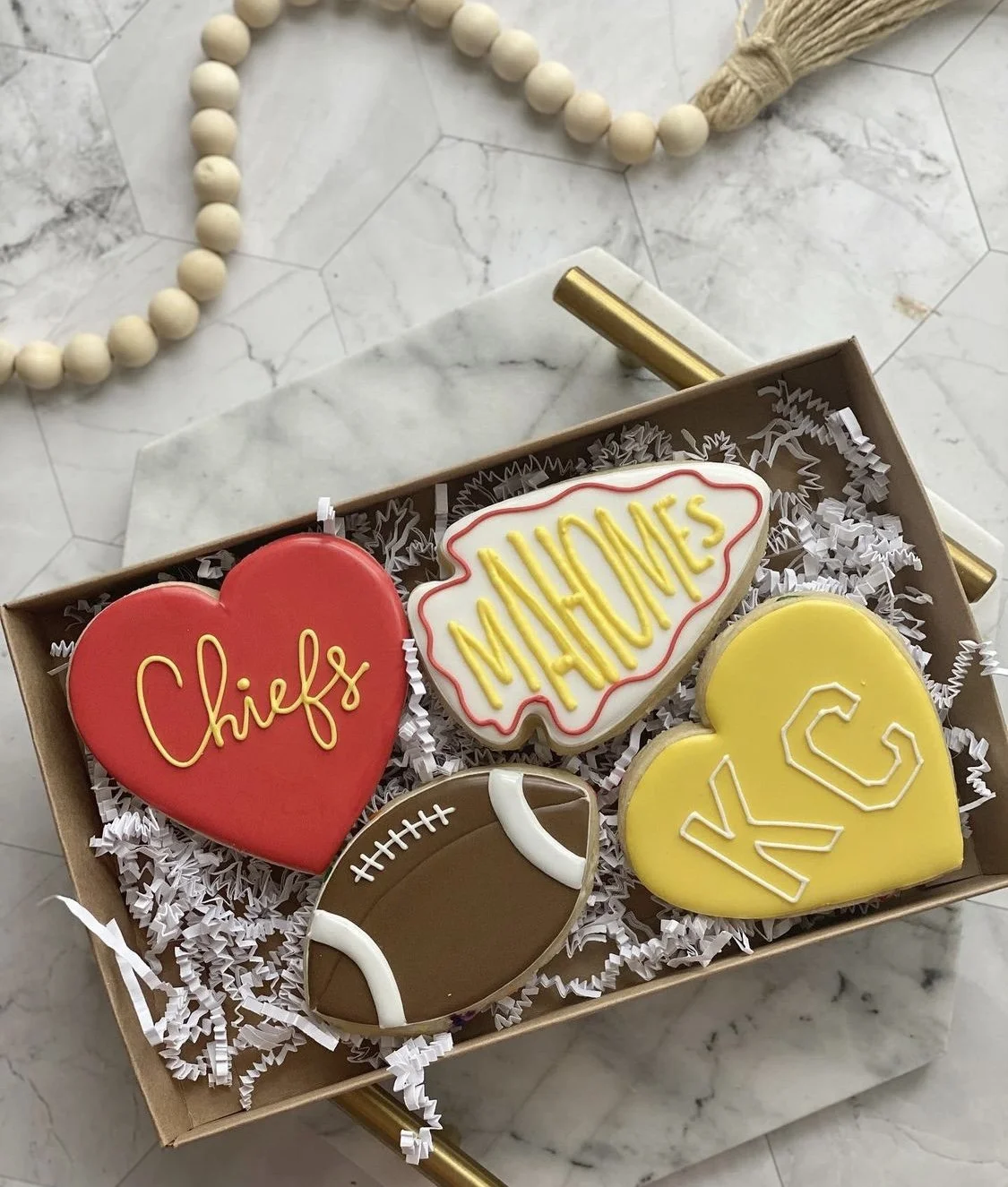 A gift box containing decorated cookies with a football and themes for Chiefs, Mahomes, and Kansas City, surrounded by gold and white beads on a marble surface.