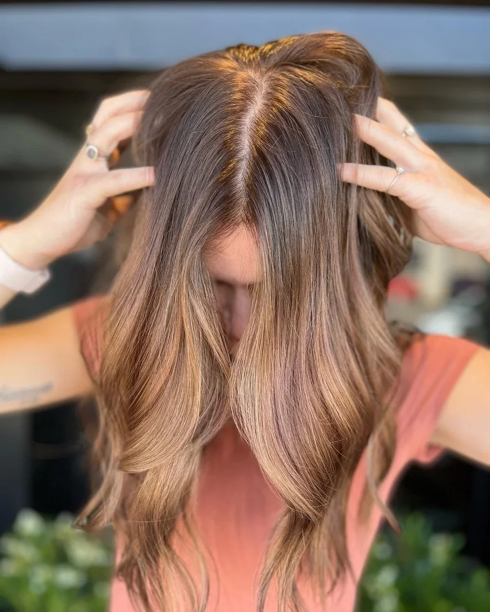 A woman with long, wavy, light brown hair. She is holding her hair with both hands, covering her face.