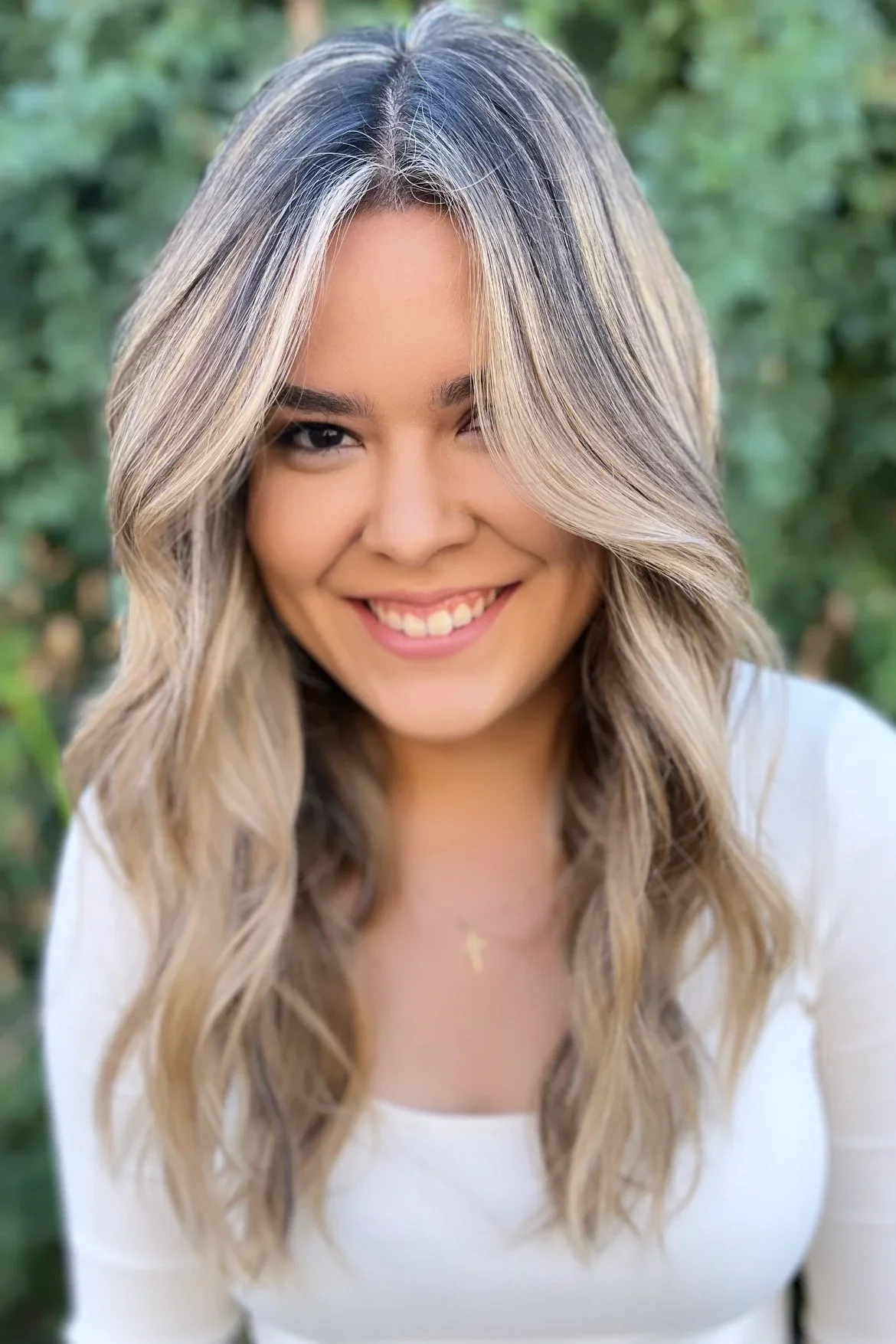 A young woman with blonde hair styled in loose waves, smiling outdoors with greenery in the background.