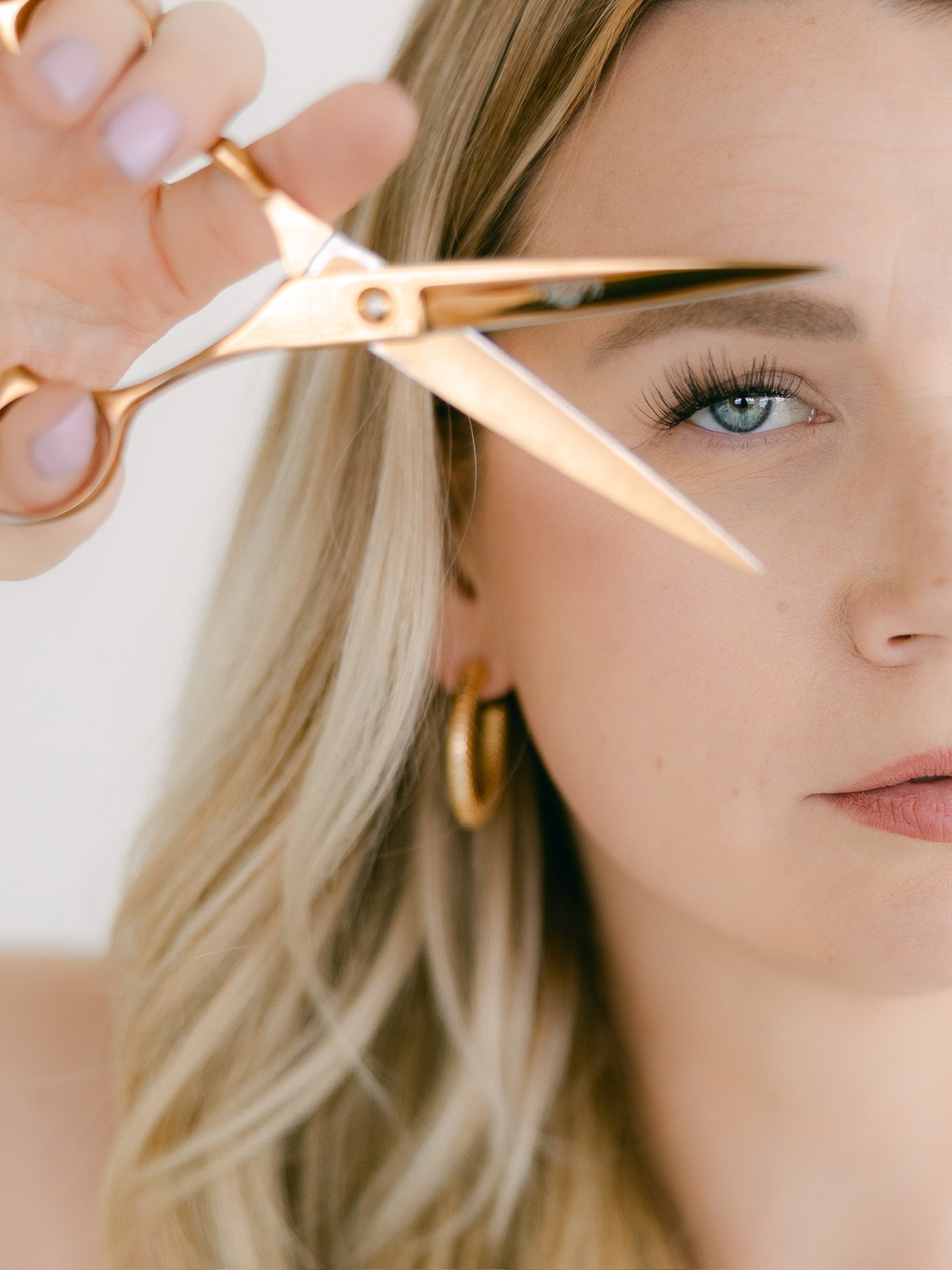 A woman with blonde hair and blue eyes is using scissors to cut her hair.