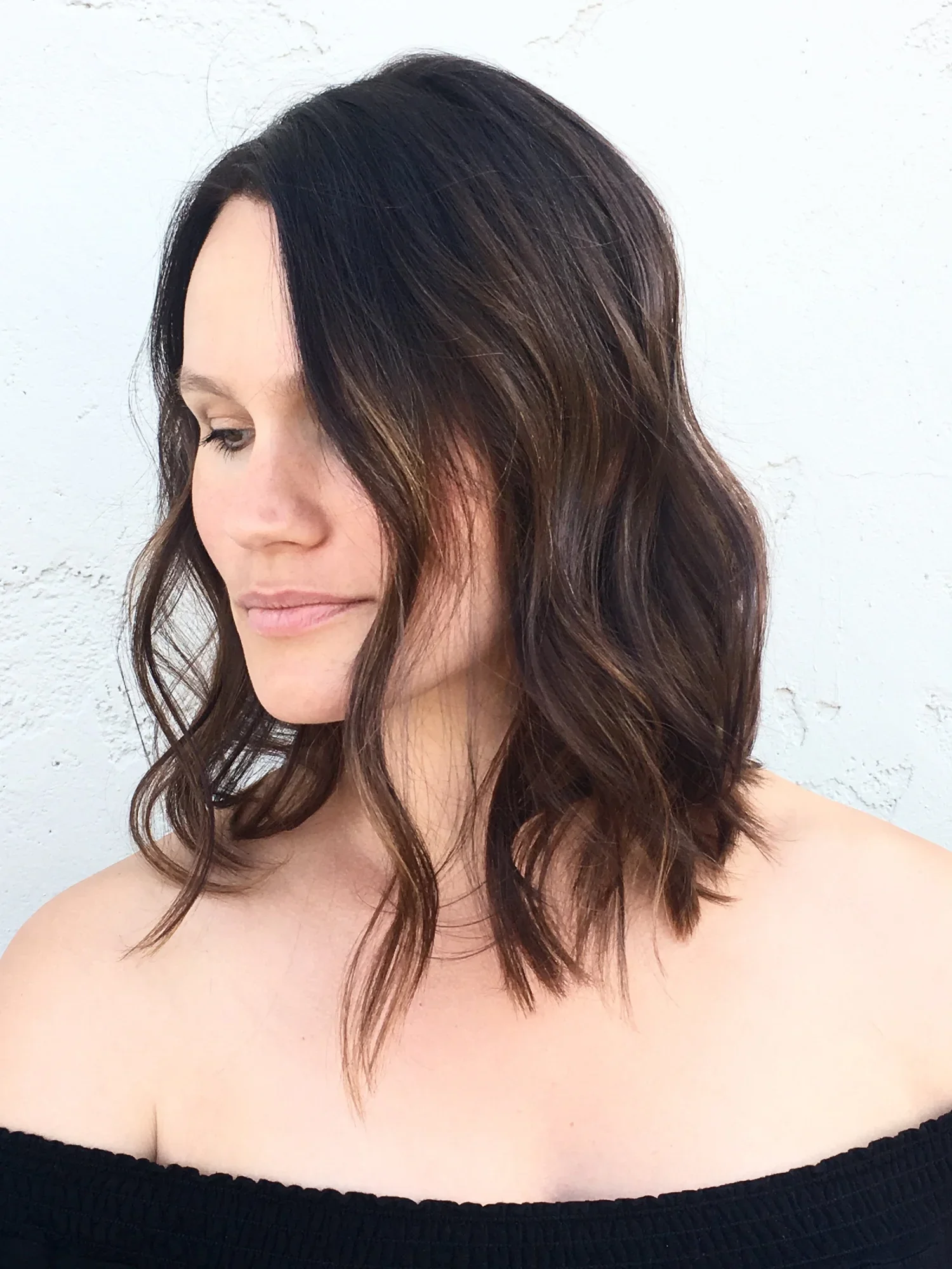 A woman with shoulder-length wavy brown hair with highlights, wearing an off-the-shoulder black top, standing against a white textured wall.