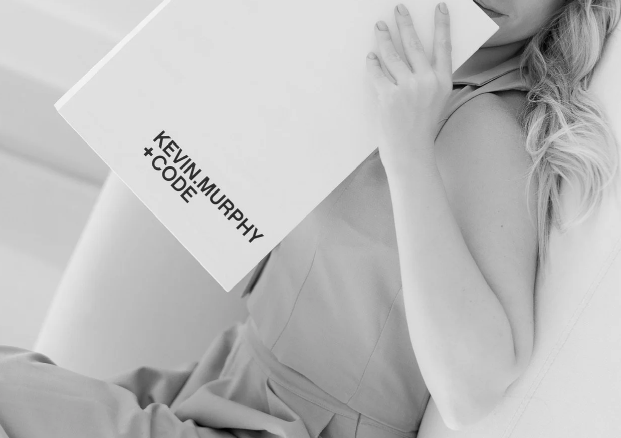 A woman with long, wavy hair is holding a white piece of paper with text on it and is sitting on a couch. The text on the paper reads "KEVIN.MURPHY + CODE." The image is in black and white.