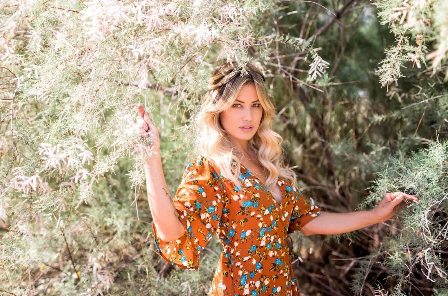 A woman with blonde, wavy hair stands among tall, spiky desert plants, wearing an orange floral dress, looking at the camera.