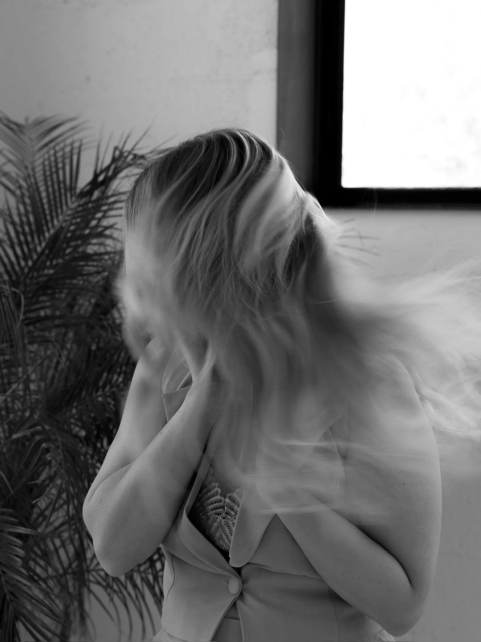 Black and white photo of a woman with long hair, blurred motion, sitting near a window, with a plant beside her.