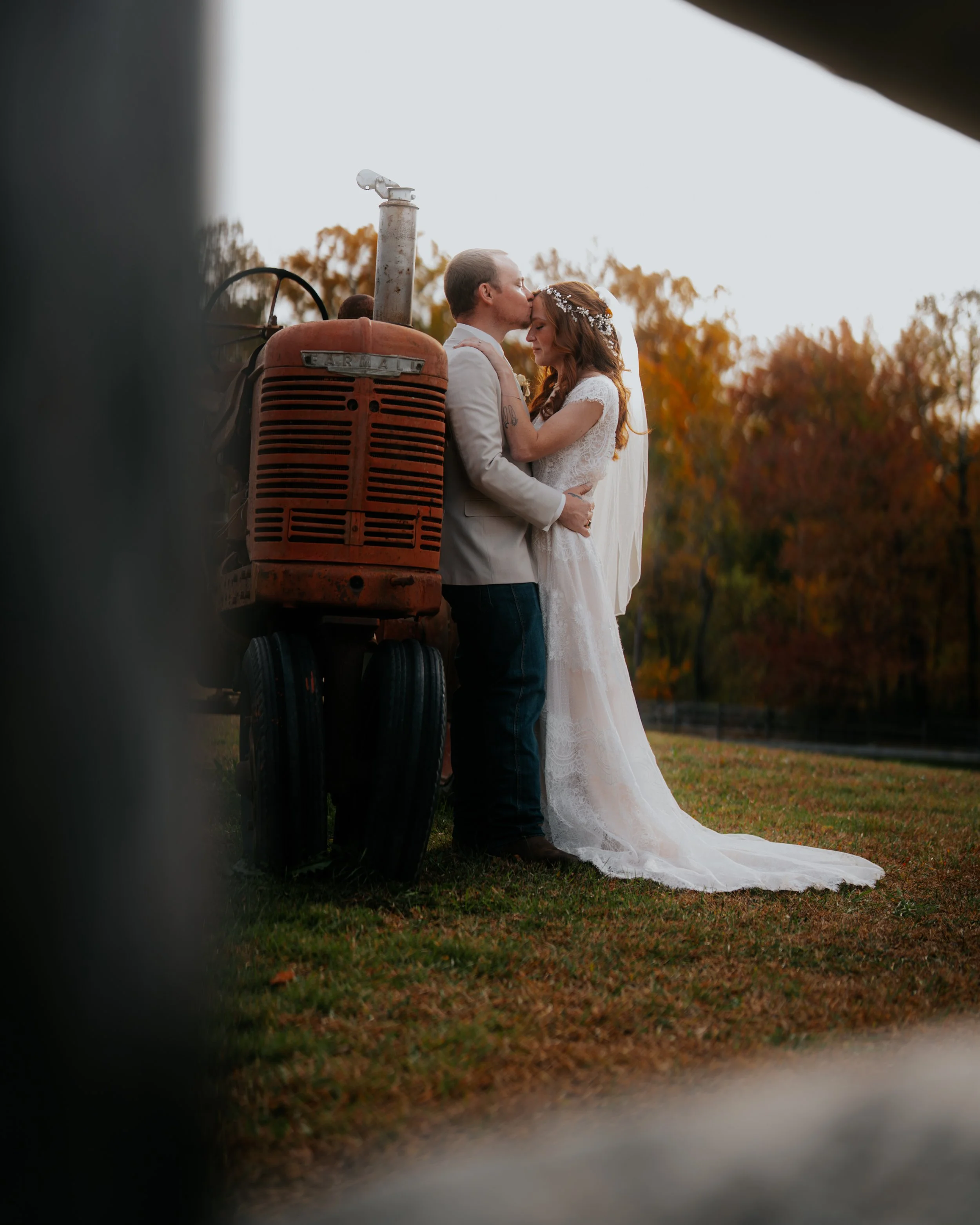 A newlywed couple in wedding attire sharing a kiss outdoors, with autumn trees in the background and a vintage tractor nearby.