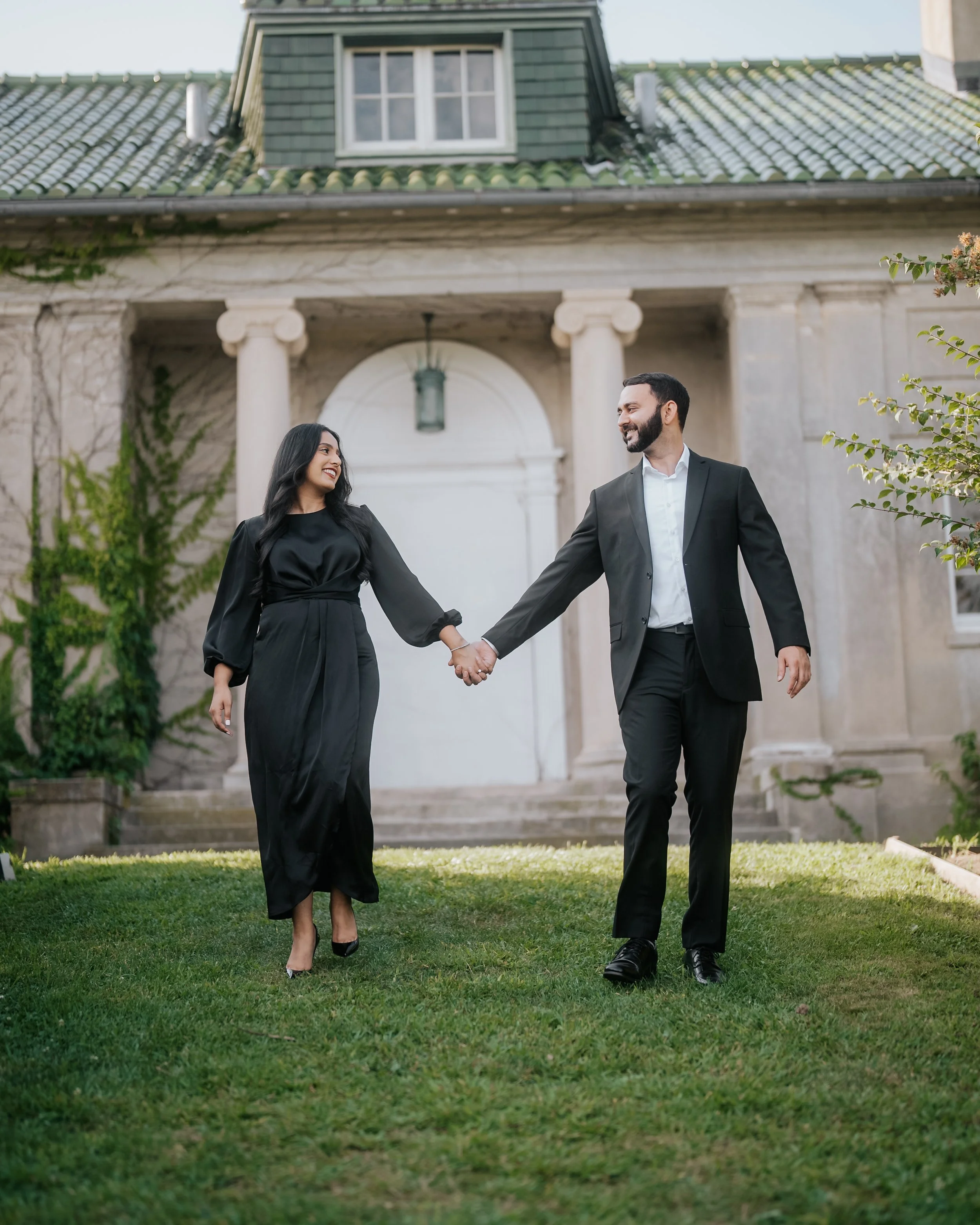 A man and woman in formal black attire holding hands, walking in front of a large house with columns and a green tiled roof.