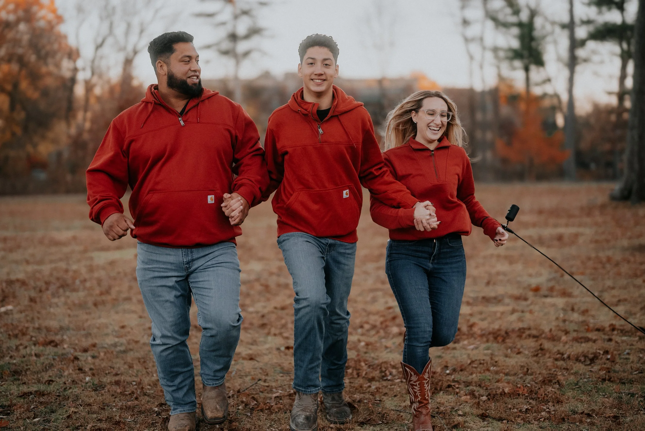 Three people walking outdoors in autumn, wearing matching red hoodies, smiling and holding hands. One person is holding a selfie stick. There are trees and fallen leaves in the background.