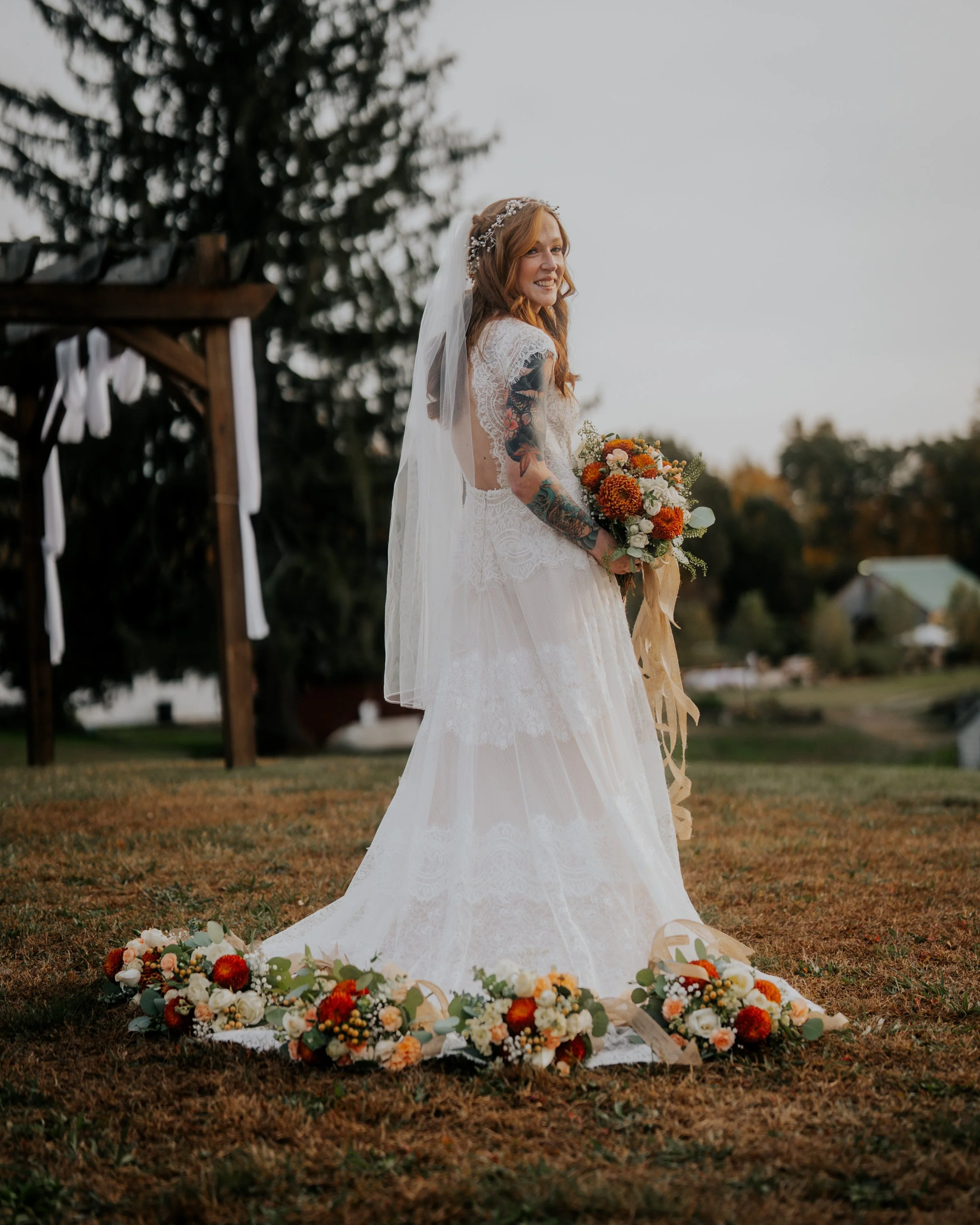 Bride in a lace wedding dress holding a bouquet of orange, white, and yellow flowers, standing on a grassy field with floral arrangements at her feet, during a wedding ceremony outdoors at dusk.