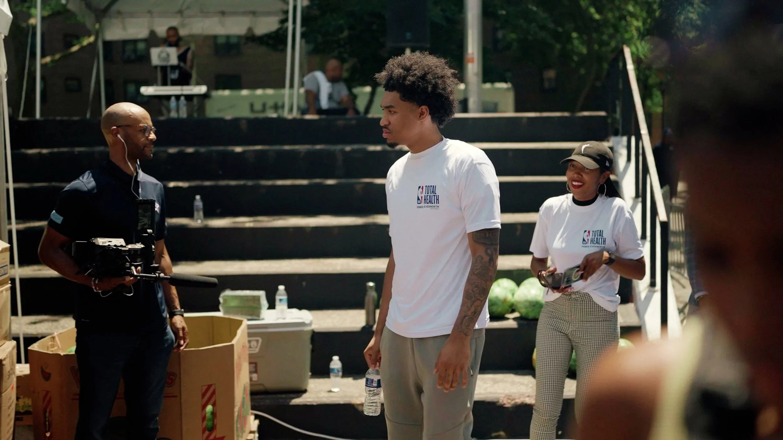 People standing outdoors at an event, with a man holding a camera on a stabilizer, a woman holding a microphone and a water bottle, and a young man with curly hair and tattoos wearing a white 'Total Health' T-shirt in the foreground.