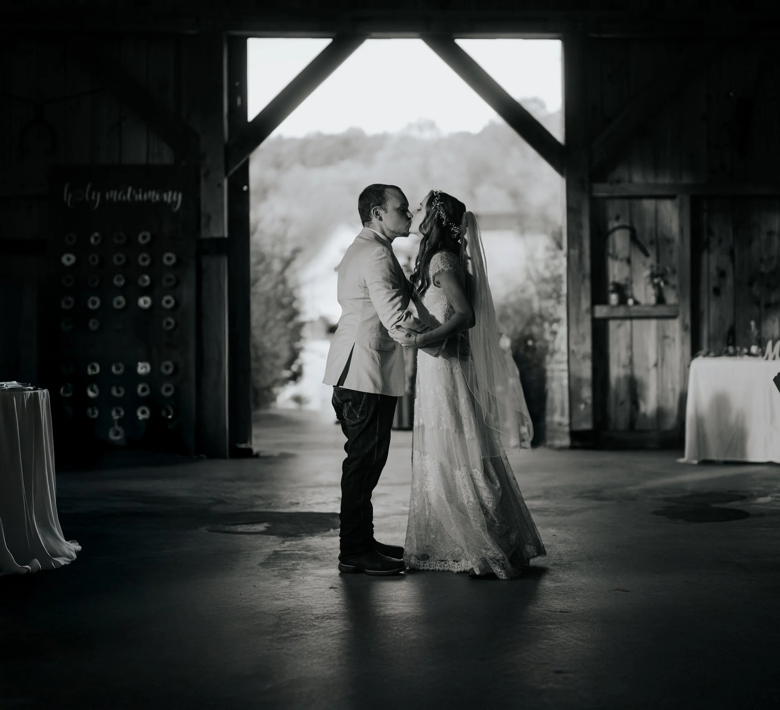 A black-and-white photo of a bride and groom kissing inside a rustic barn with open doors, natural light streaming in.
