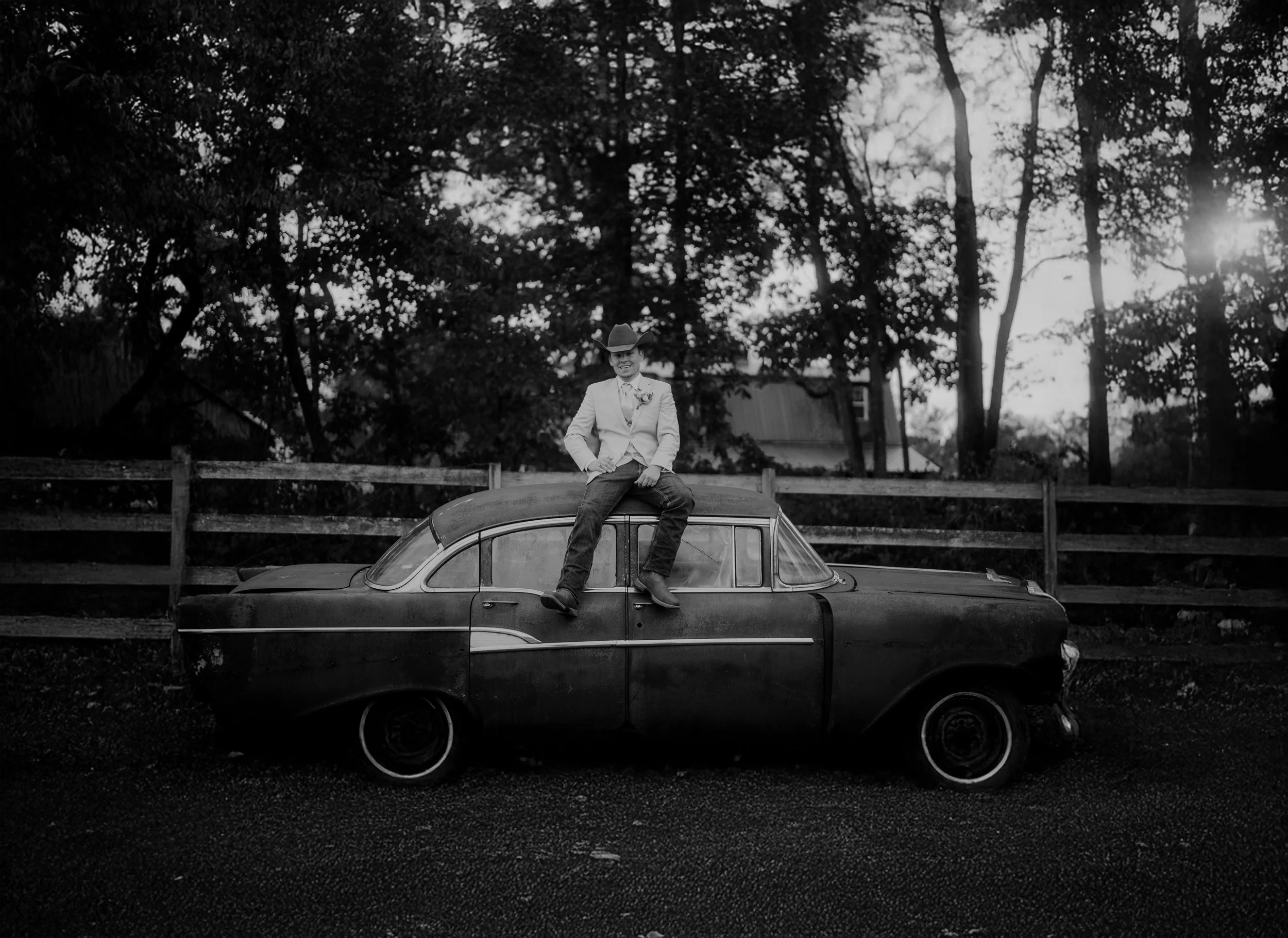 Black and white photo of a young man sitting on top of a vintage car, wearing a cowboy hat, suit jacket, and jeans, with trees and a wooden fence in the background during sunset.
