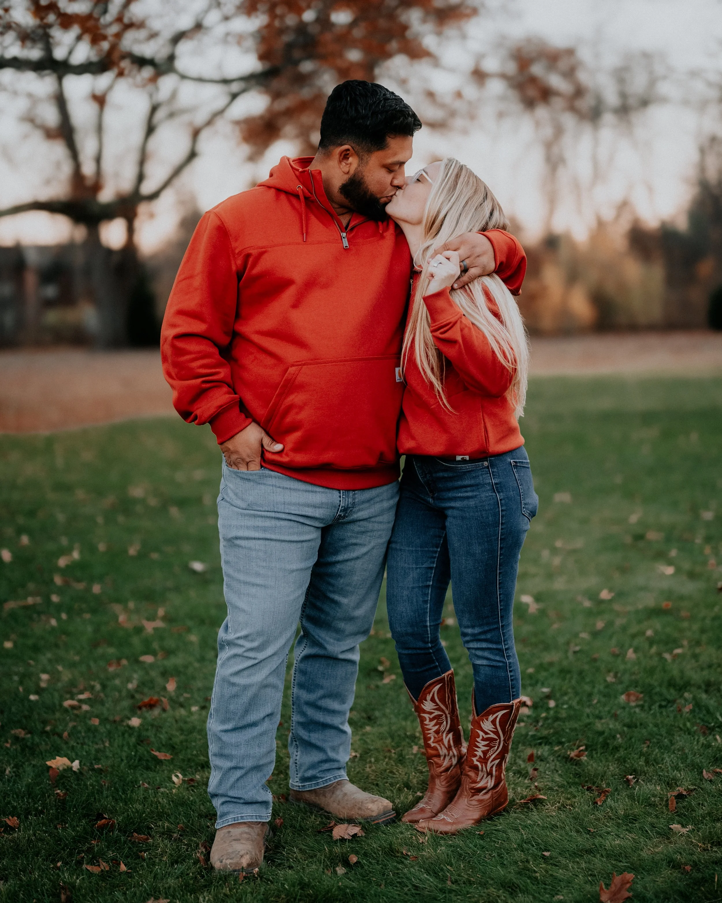 A couple in red hoodies sharing a kiss outdoors in a park during fall, with trees and grass in the background.