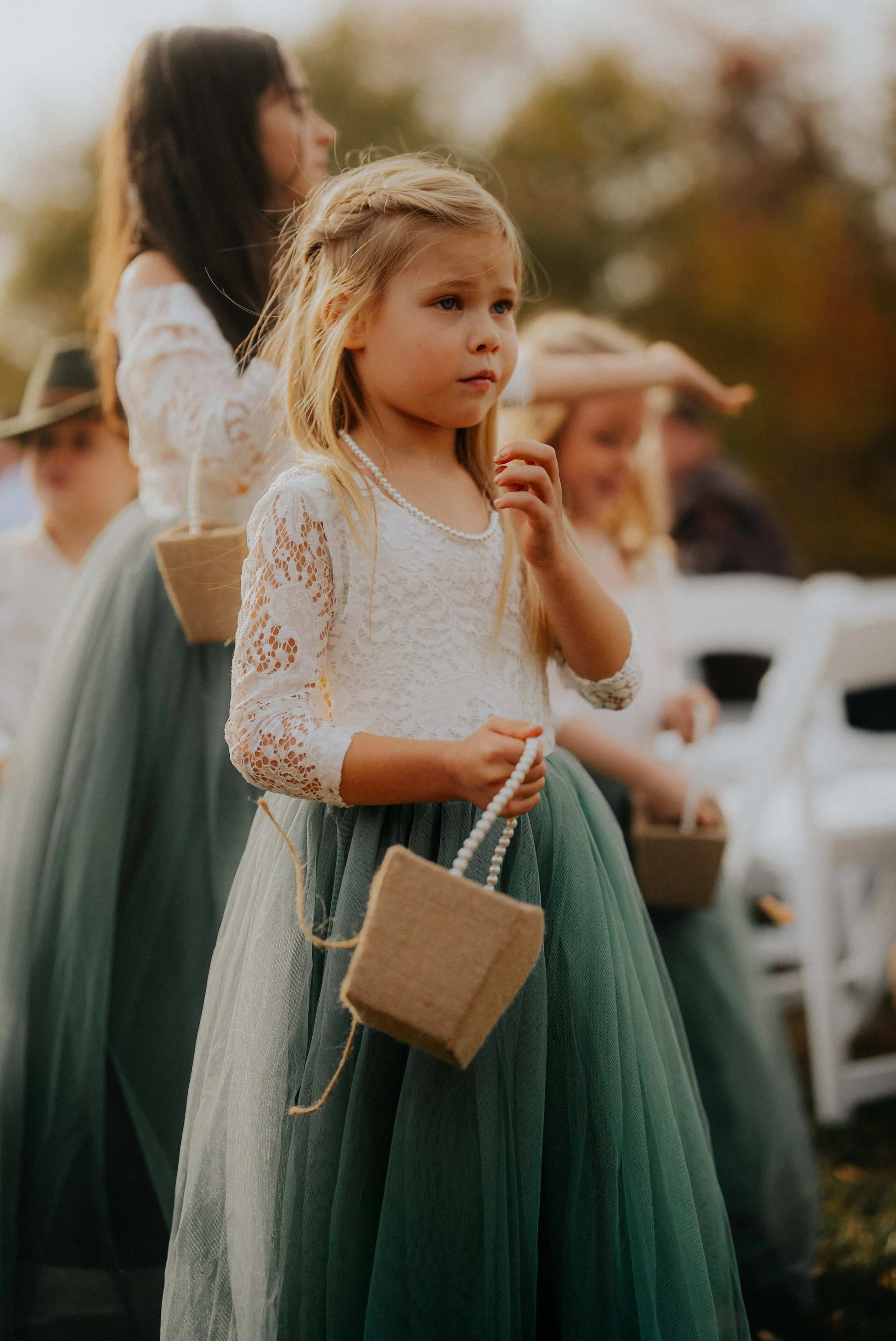 Young girl holding a small woven basket, wearing a white lace top and teal tulle skirt, during an outdoor event with others in similar attire.