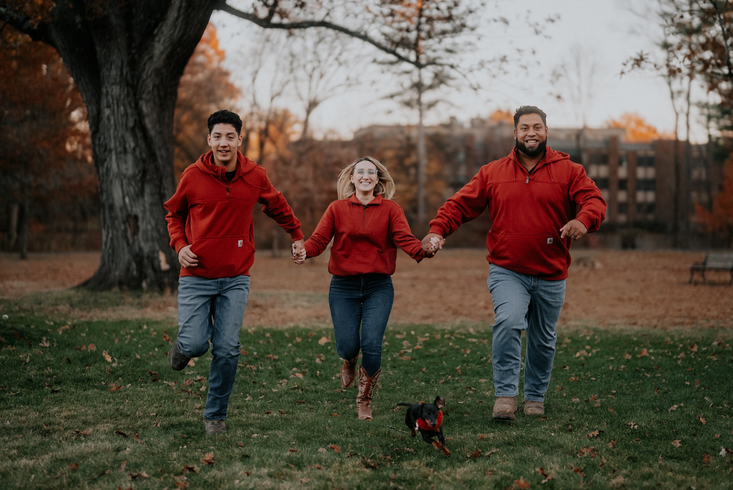 Three people running and holding hands in a park during fall with a small dog in front, all wearing red jackets.