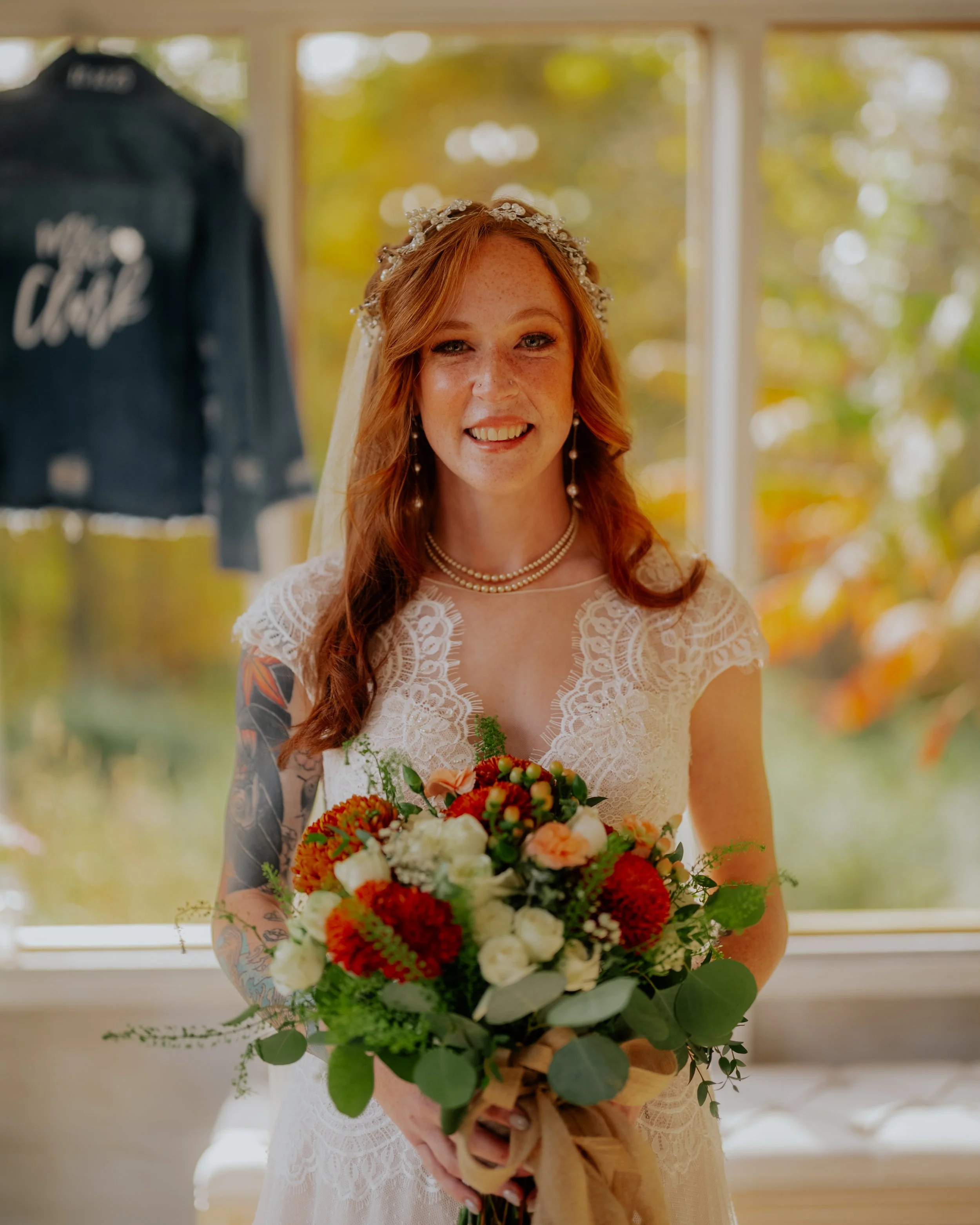 A smiling bride with red hair and tattoos on her arm, wearing a lace wedding dress, pearl jewelry, and a floral headpiece, holding a bouquet of white, red, and orange flowers, standing indoors with a large window and autumn foliage outside.