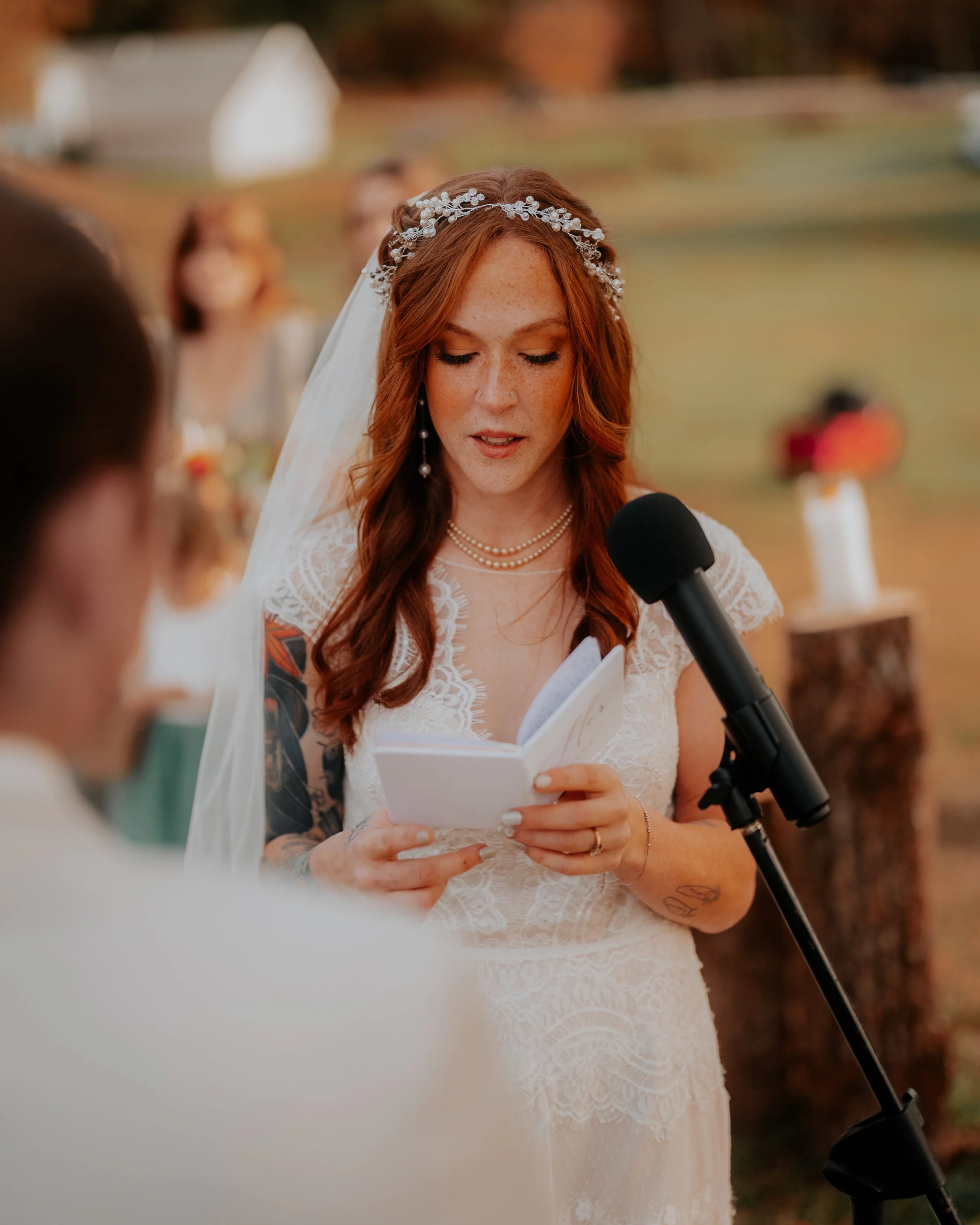 A woman with long red hair, tattoos, and wearing a white lace wedding dress reads vows from a small book during an outdoor wedding ceremony, with a black microphone in front of her.