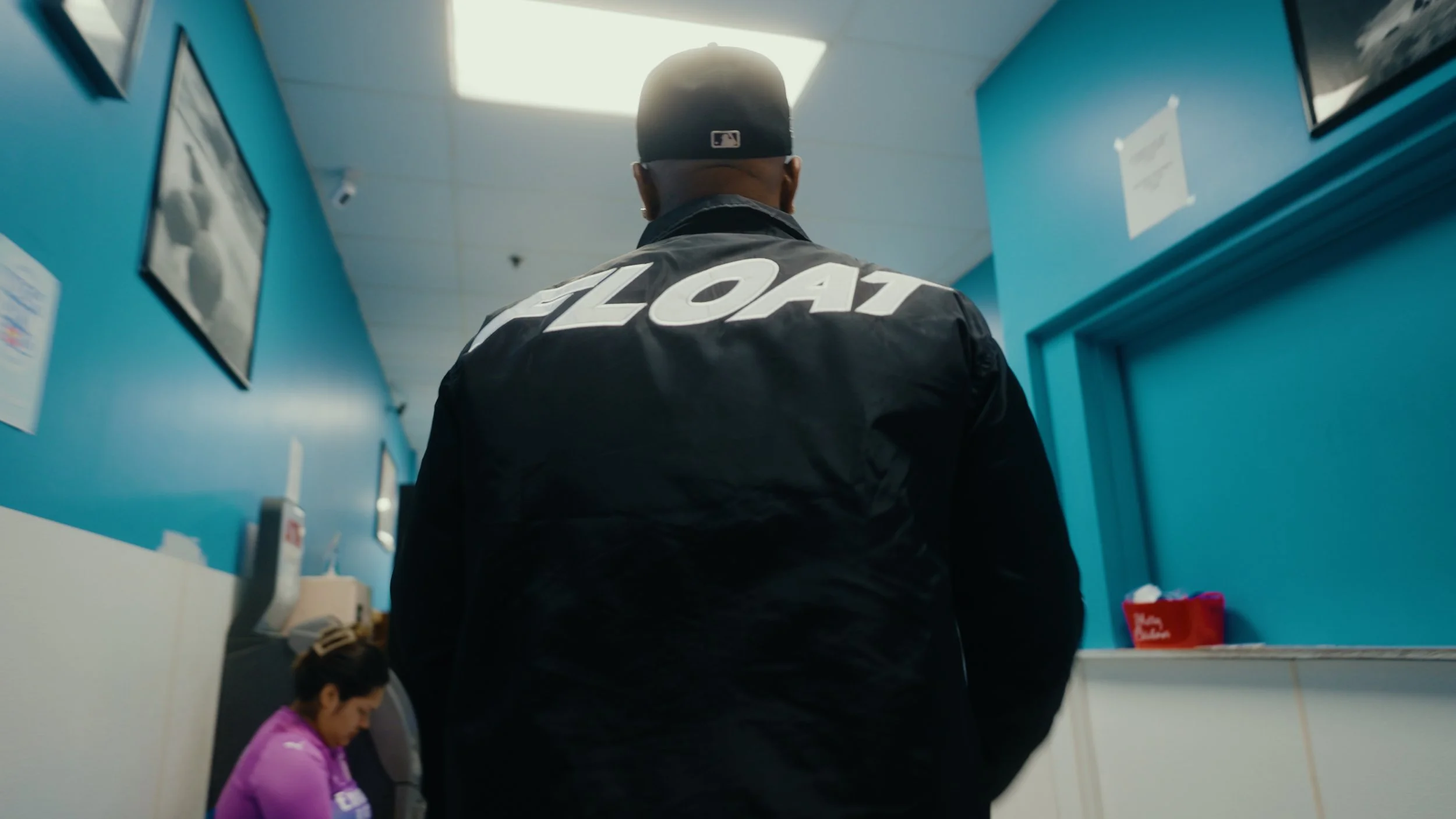 A person wearing a black jacket with the word 'FLOAT' on the back, standing in a blue wall corridor, with a woman in a purple shirt sitting against the wall in the background.